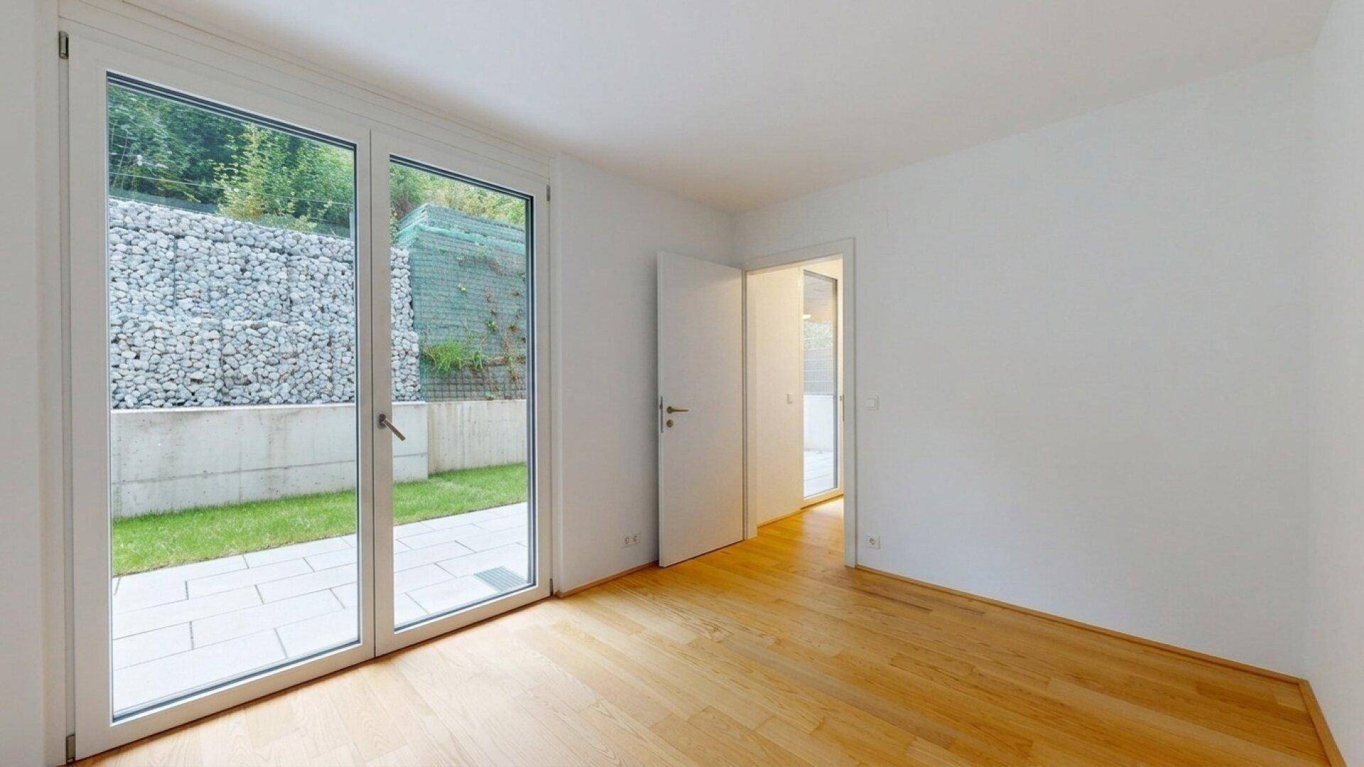Leeres Zimmer mit Holzboden und Zugang zu einem Außenbereich mit Gabionenwand und Rasen.