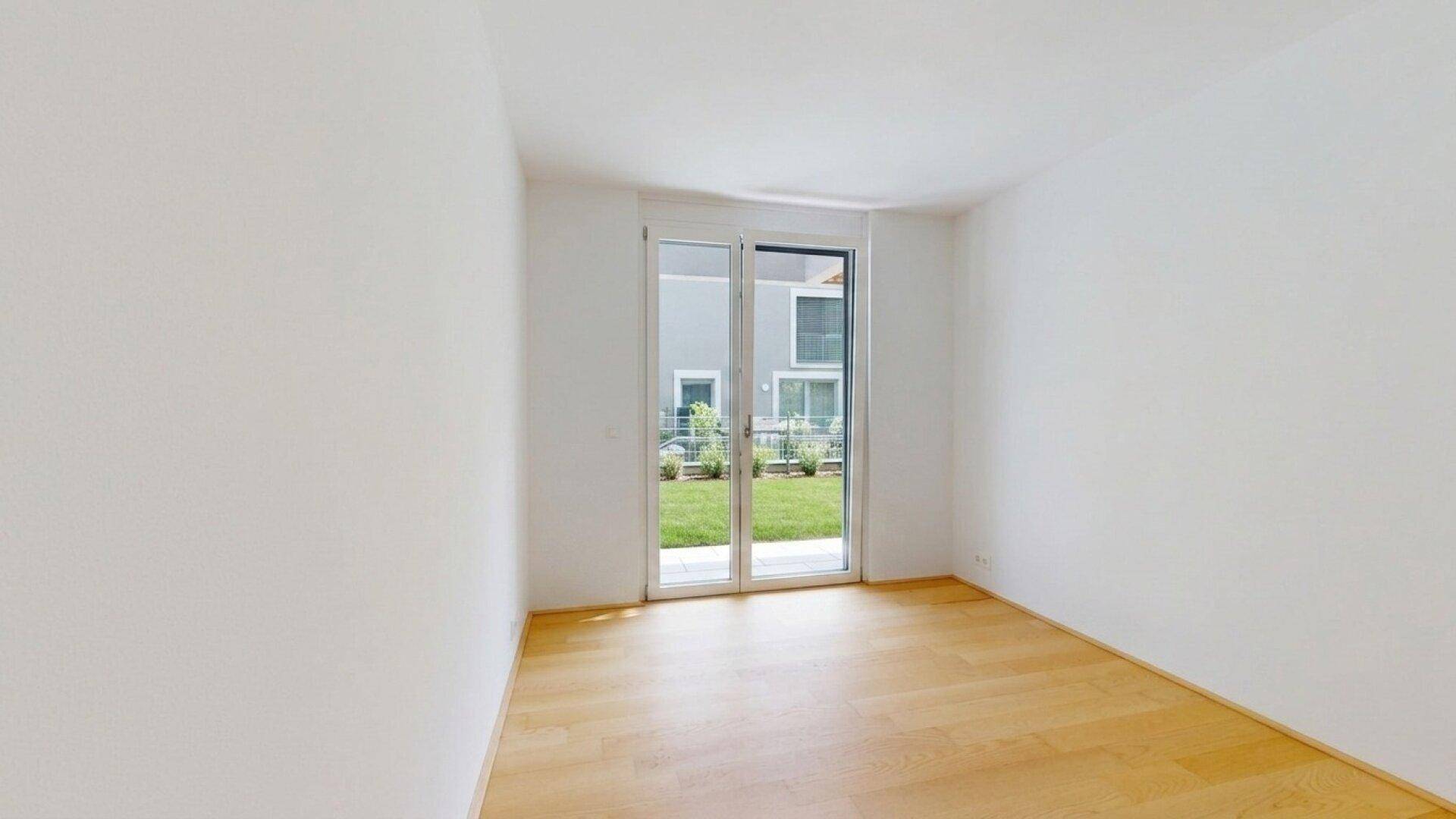 Leeres Zimmer mit Holzboden und weißen Wänden, mit Blick auf die grüne Umgebung.