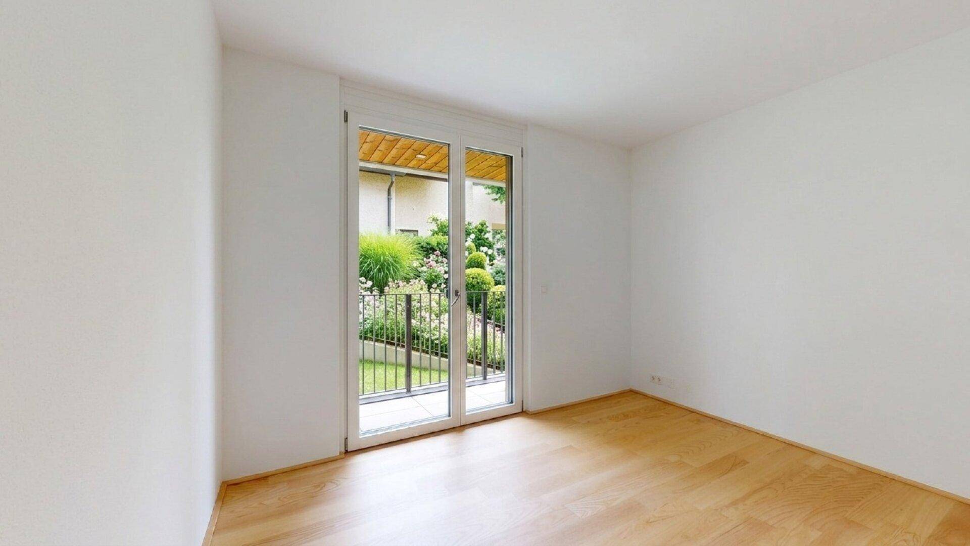 Helles Zimmer mit Holzboden und Zugang zu einem kleinen, bepflanzten Gartenbereich.