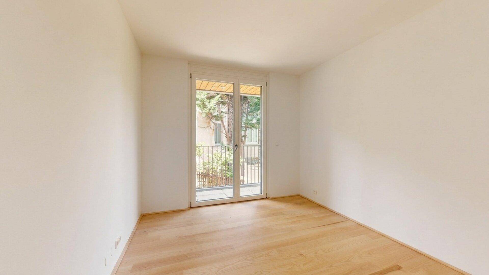 Helles Zimmer mit Holzboden und Zugang zu einem kleinen Balkon, umgeben von Grün.