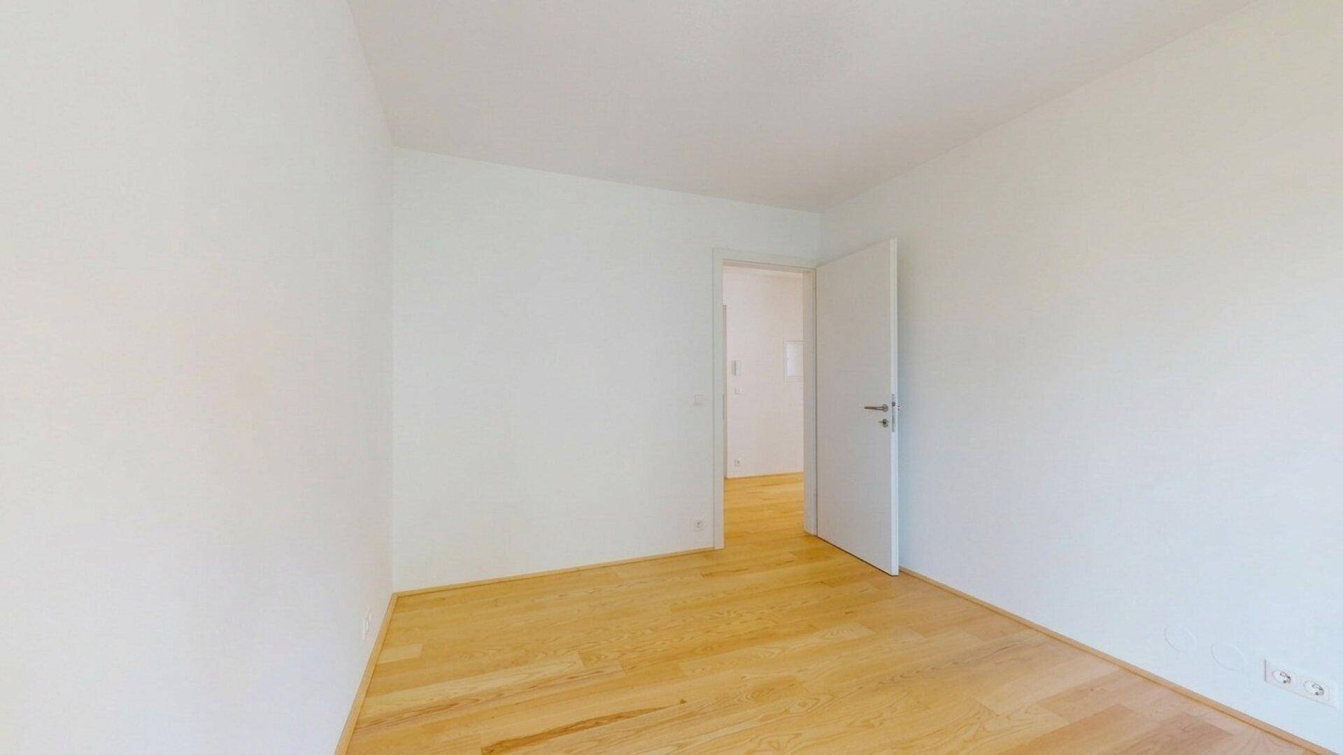 Leeres Zimmer mit hellem Holzboden und einer offenen Tür, die in einen weiteren Raum führt.