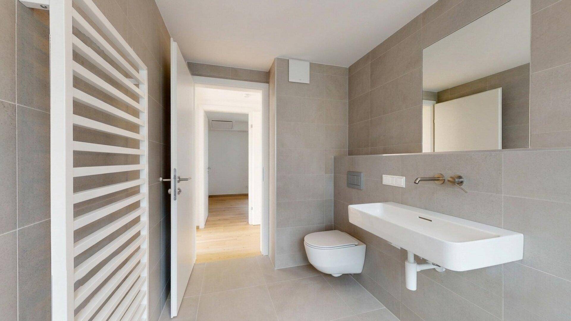Modernes Badezimmer mit Wand-WC, Waschtisch und großem Spiegel, alles in hellgrauen Fliesen.