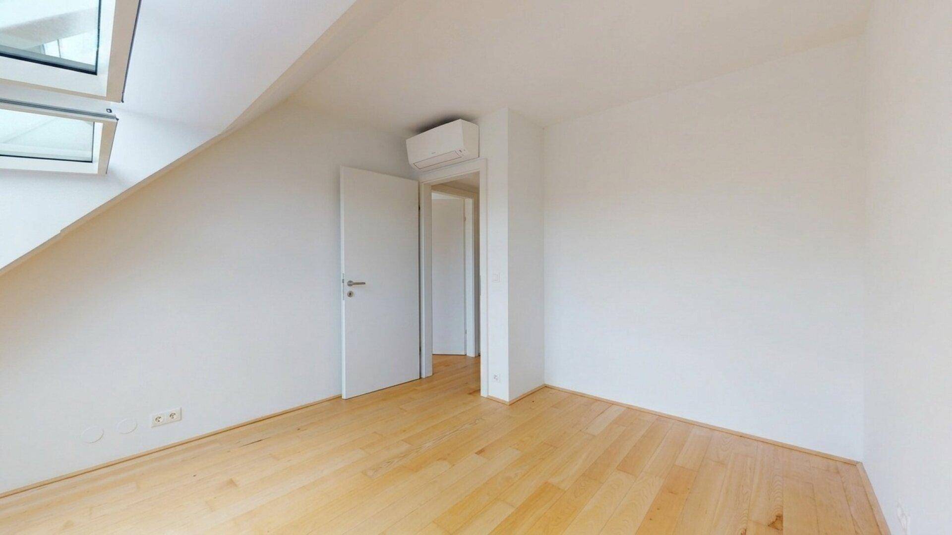 Geräumiges Zimmer mit Holzboden, weiß gestrichenen Wänden und einer Klimaanlage.