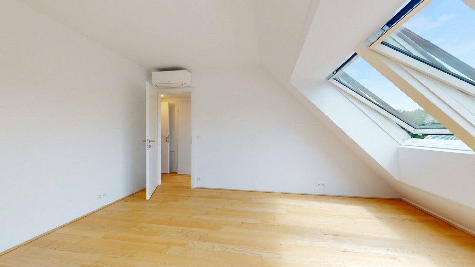 Heller Raum mit Holzboden, schrägen Wänden und einem Dachfenster, das viel Licht spendet.