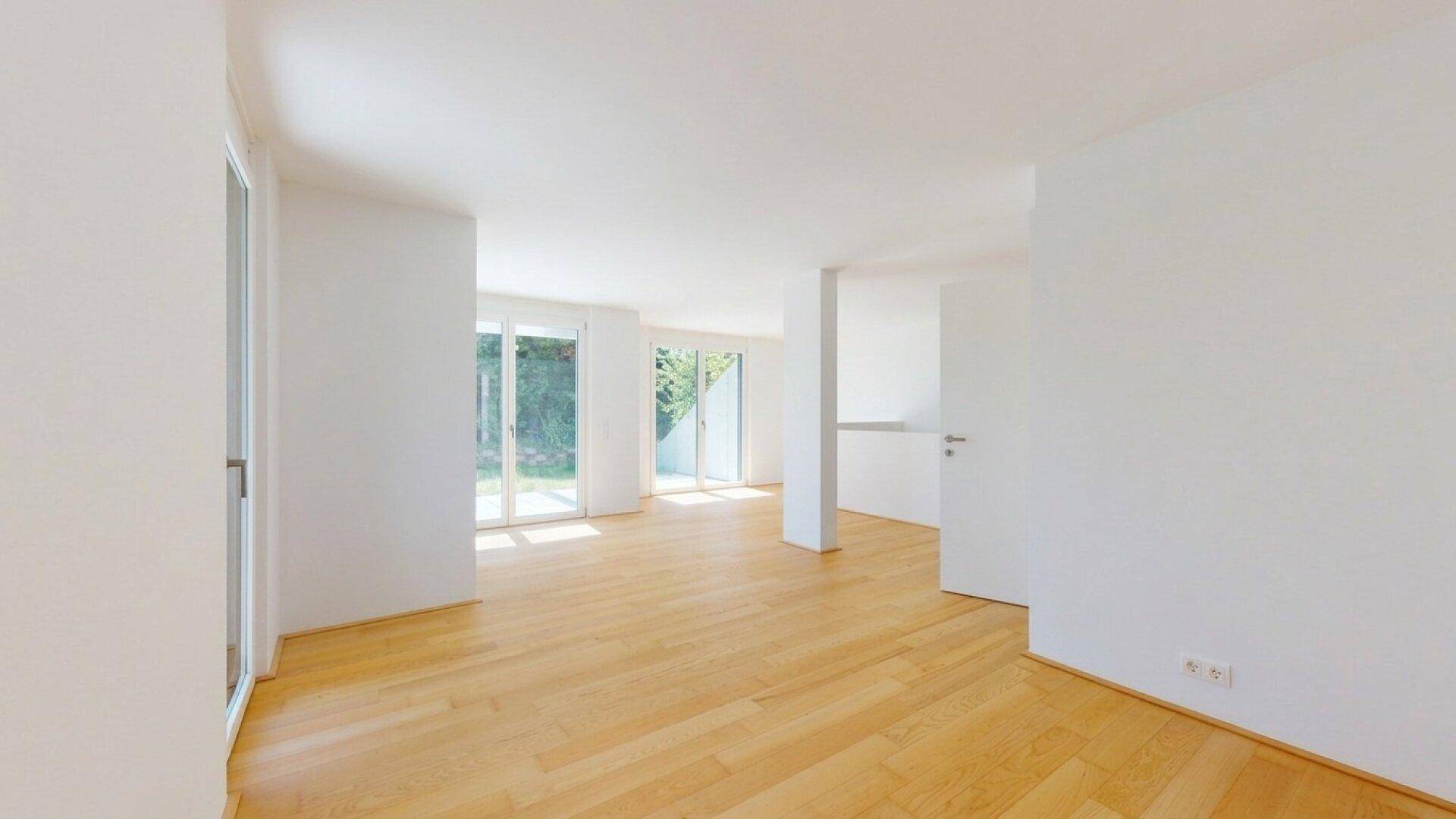 Heller, leerer Wohnbereich mit Holzboden und großen Fenstern zum Garten.