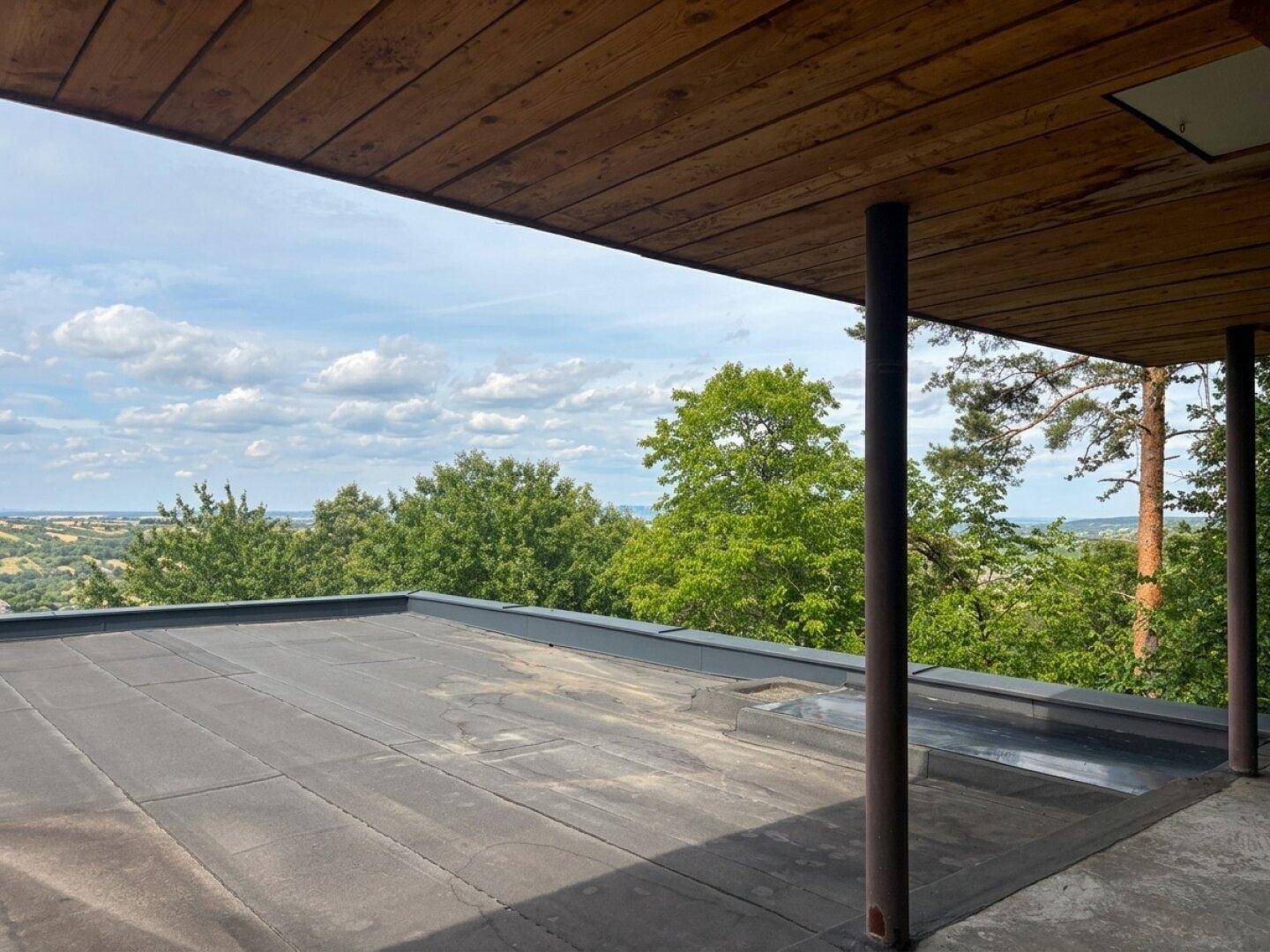 Überdachter Außenbereich mit Holzdach und weitem Ausblick auf die bewaldete Hügellandschaft.