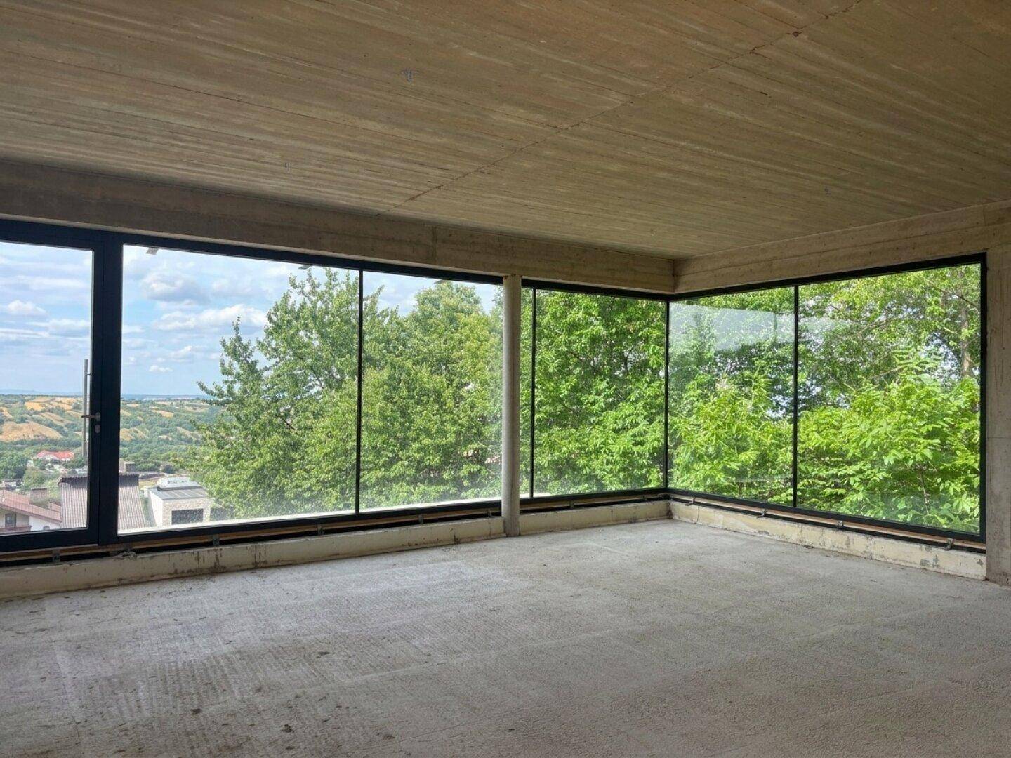 Heller Rohbau-Raum mit Eckfenstern, die einen weiten Blick auf die umliegenden Bäume bieten.