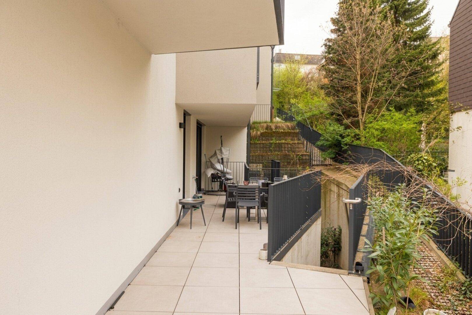 Geräumige Terrasse mit Fliesenboden und Sitzgelegenheiten, umgeben von Grün und einer Treppe.
