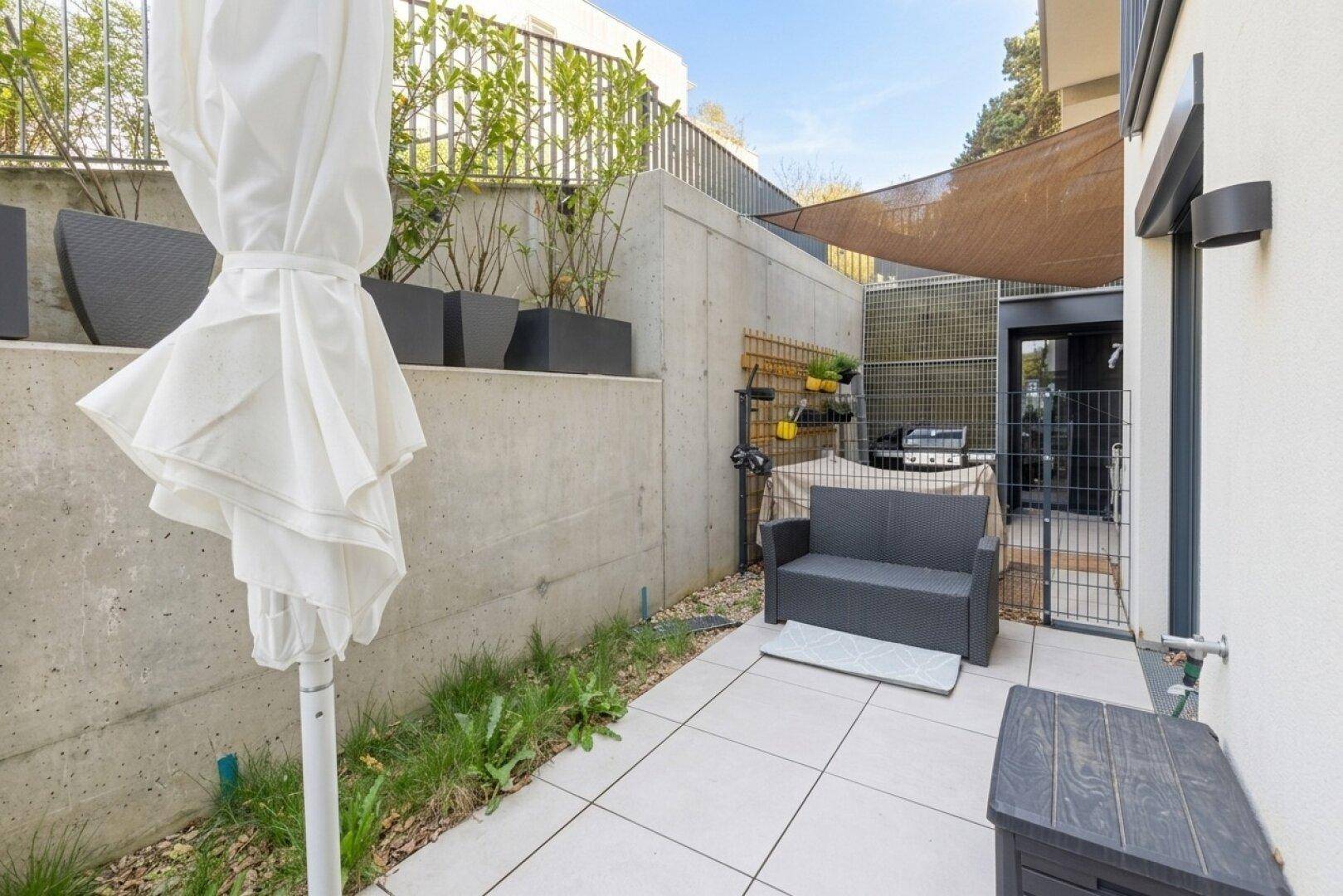 Gemütlicher Gartenbereich mit Sonnenschutz, Sitzbank und Grill, umgeben von Betonwänden.