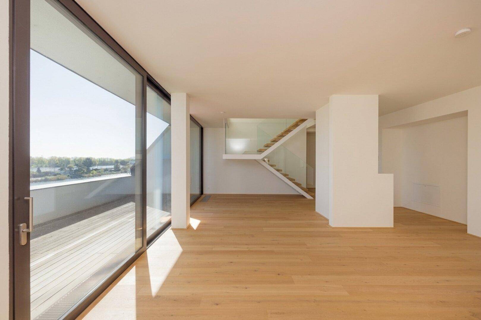 Helles, offenes Wohnzimmer mit Holzboden, großen Schiebetüren und modernem Treppenaufgang mit Glasgeländer.