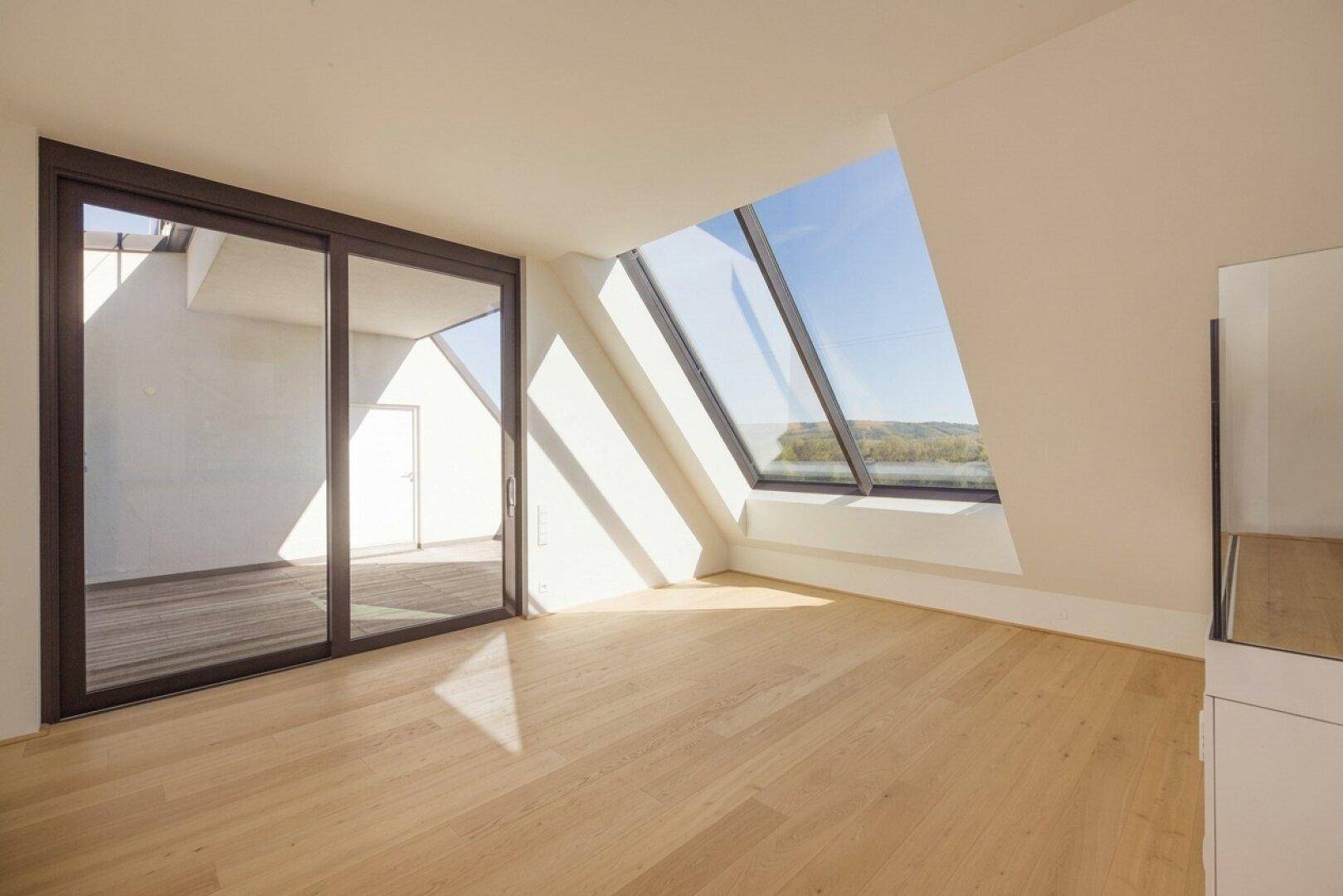 Lichtdurchfluteter Dachgeschossraum mit schrägen Wänden, Holzboden und großen, schrägen Fenstern.