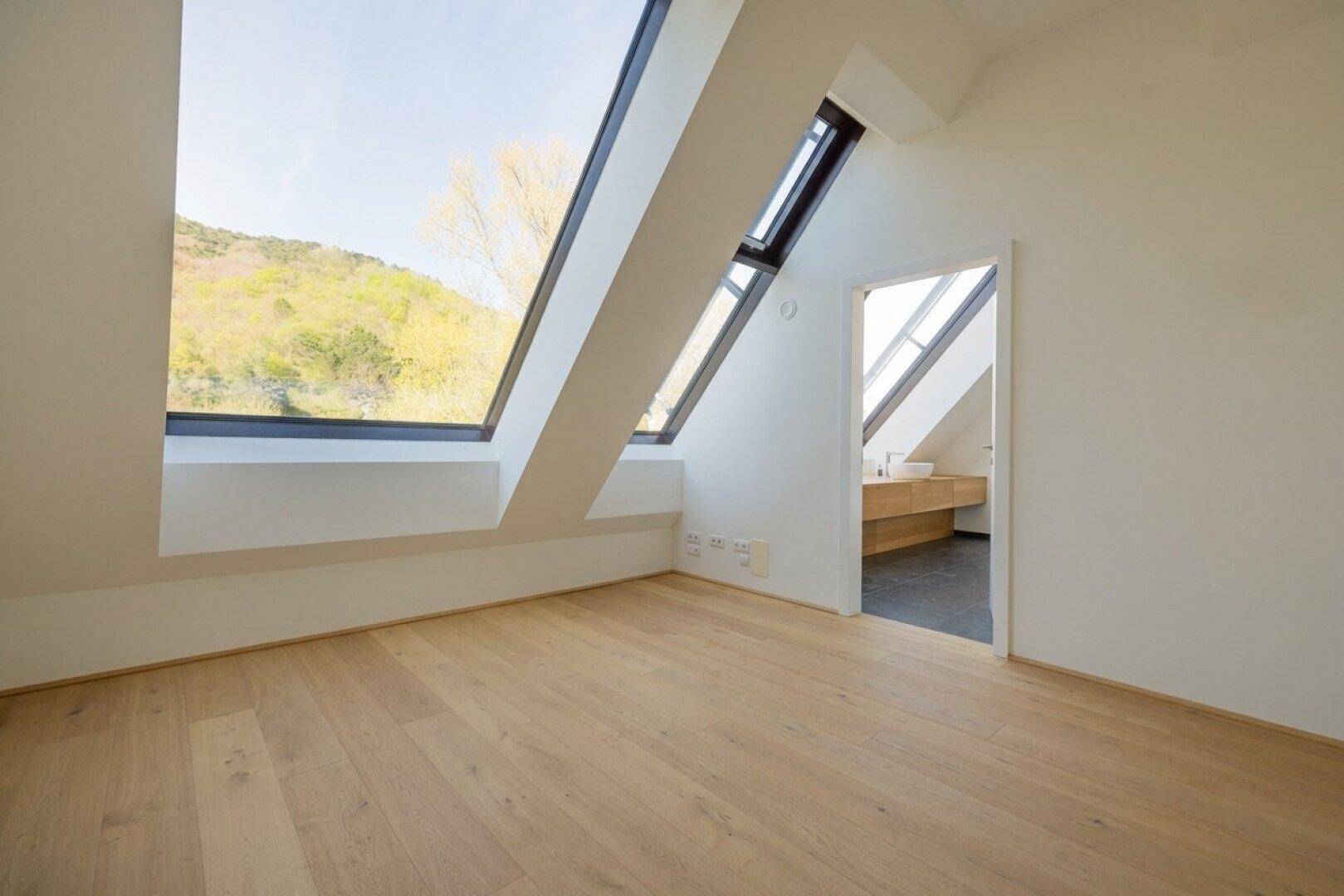 Helles Schlafzimmer im Dachgeschoss mit Holzboden, schrägen Wänden und einem großen Schrägfenster mit Aussicht.
