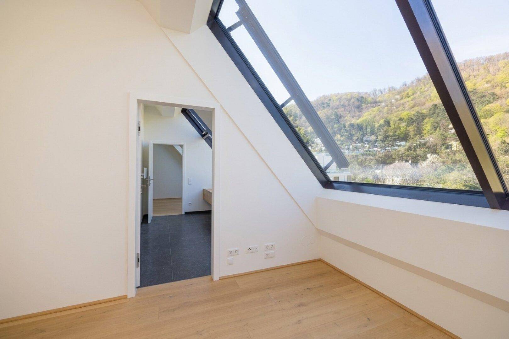 Helles Dachgeschosszimmer mit Holzboden, schrägen Wänden und einem großen Schrägfenster mit Aussicht.