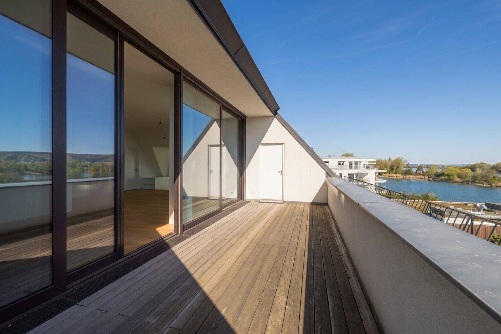 Geräumiger Balkon mit Holzdeck und weitem Blick auf den See und die grüne Landschaft unter blauem Himmel.
