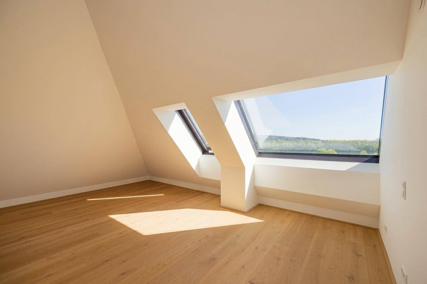 Helles Dachgeschosszimmer mit Holzboden und zwei schrägen Fenstern, die viel Sonnenlicht hereinlassen.