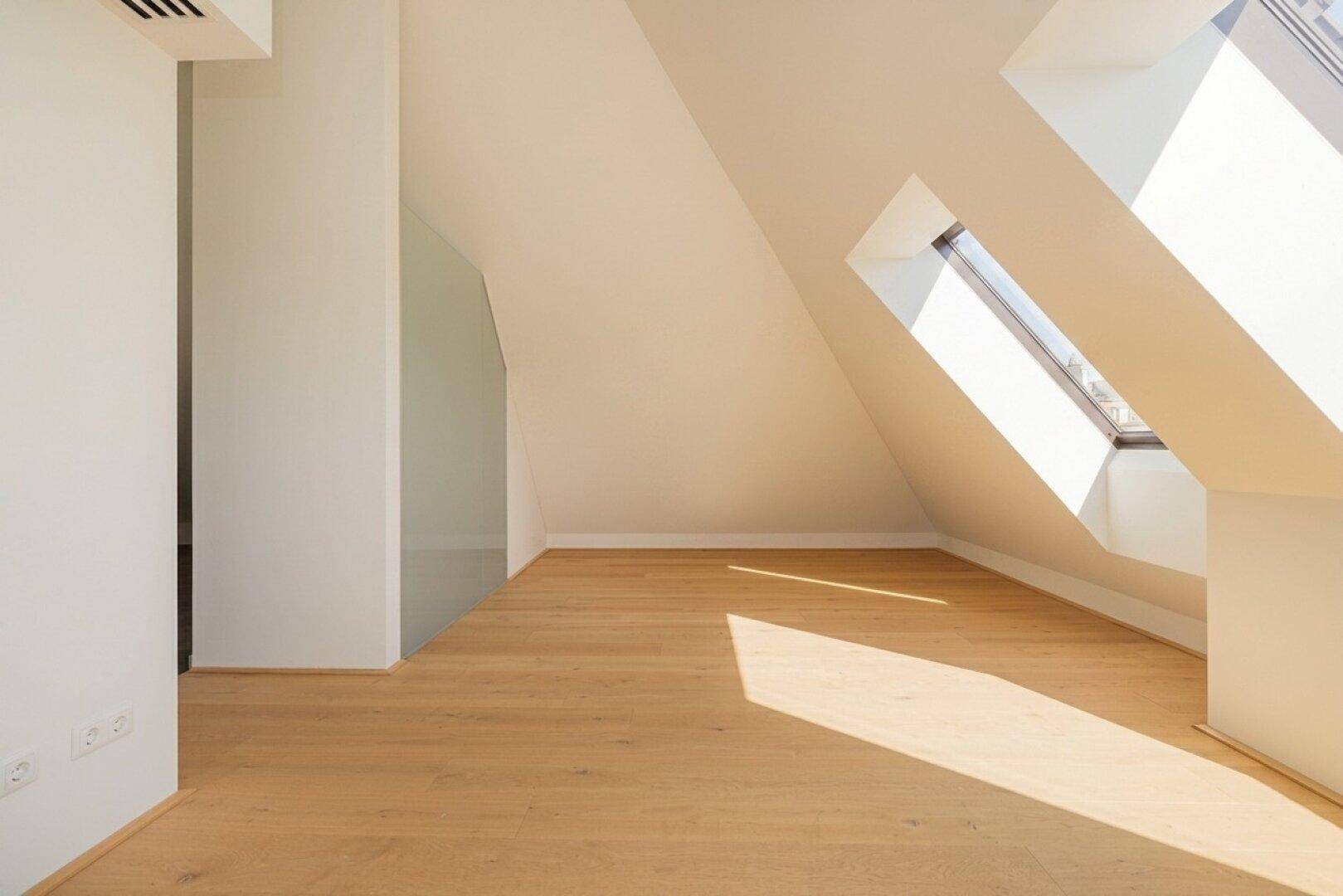 Geräumiges Dachgeschosszimmer mit Holzboden und schrägen Wänden, durchflutet von natürlichem Sonnenlicht.