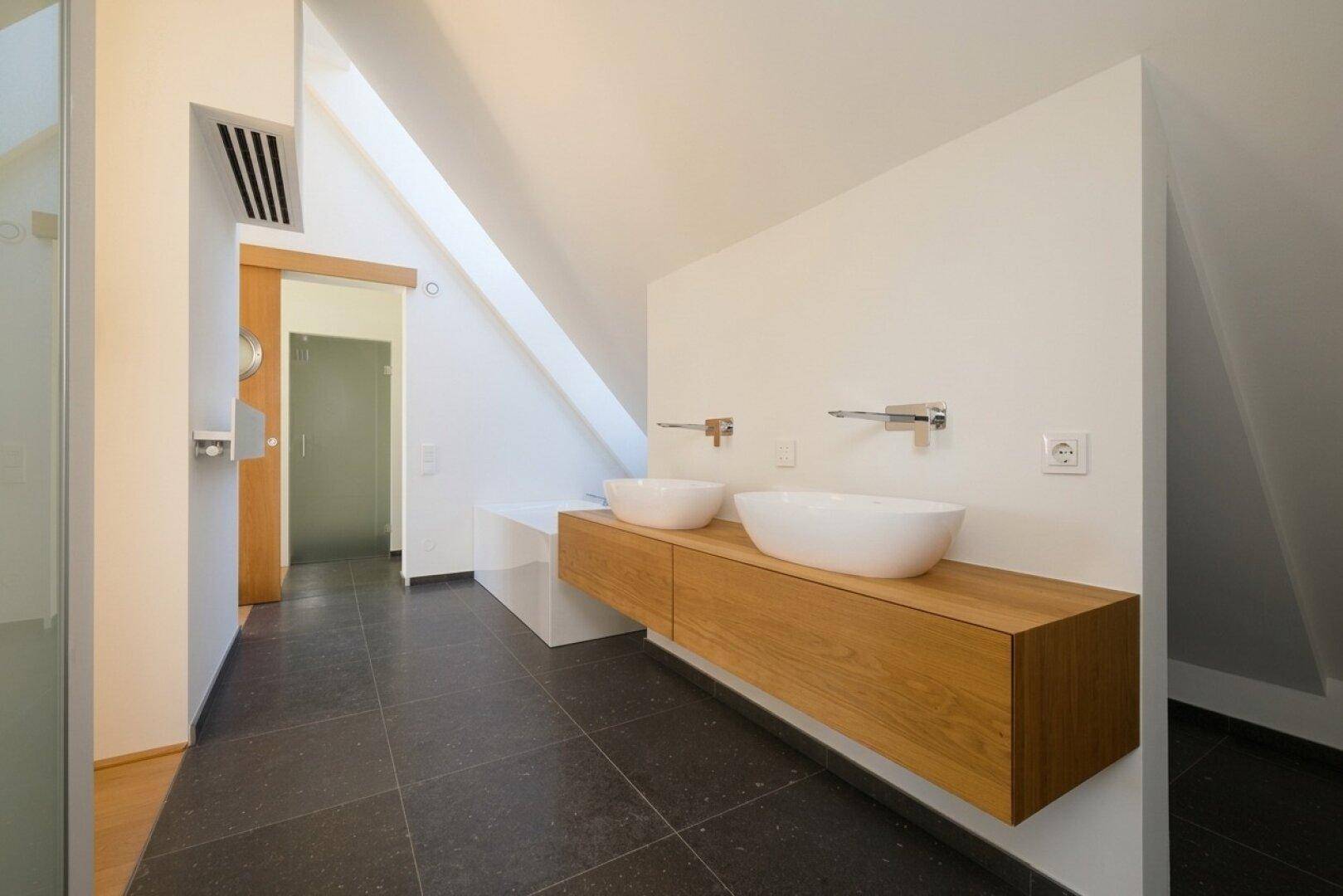 Modernes Badezimmer mit Doppelwaschbecken auf Holzunterschrank und dunklen Fliesen, hell und geräumig.