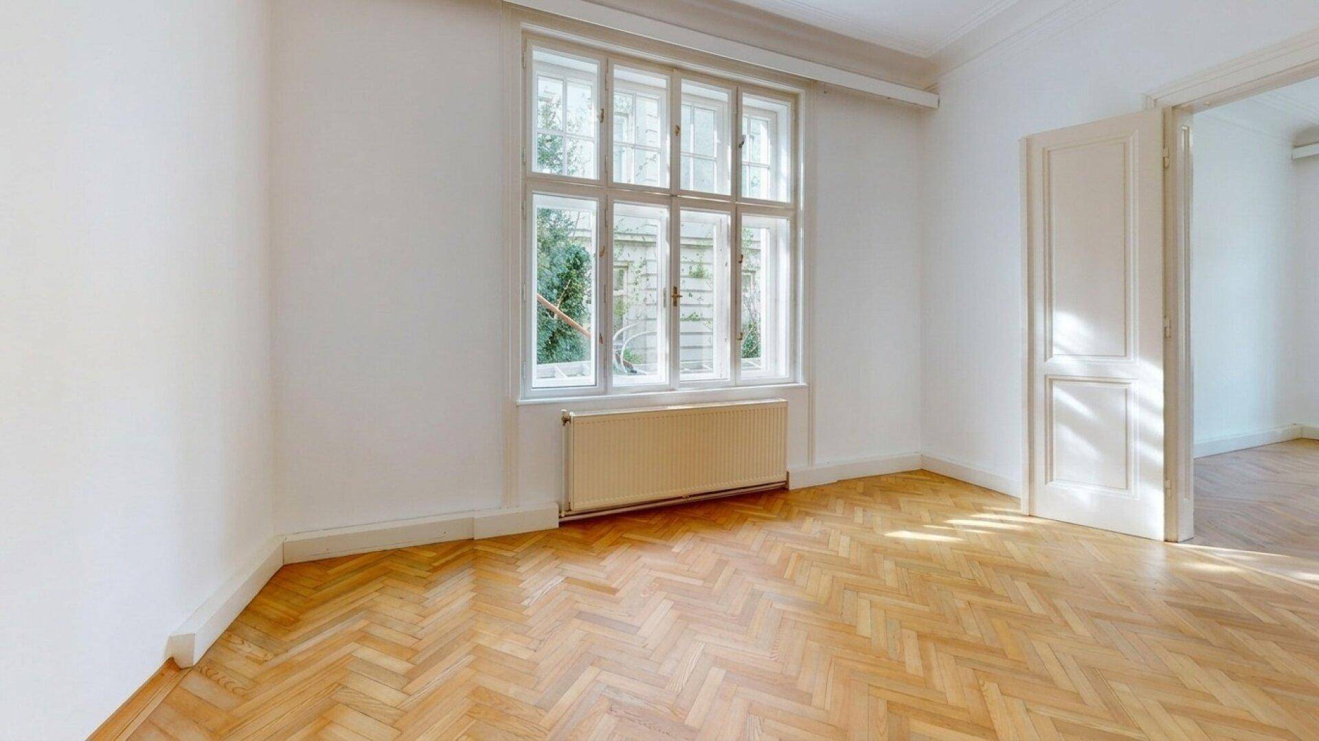 Lichtdurchflutetes Zimmer mit klassischem Fischgrätparkett und Blick ins Grüne durch große Fenster.
