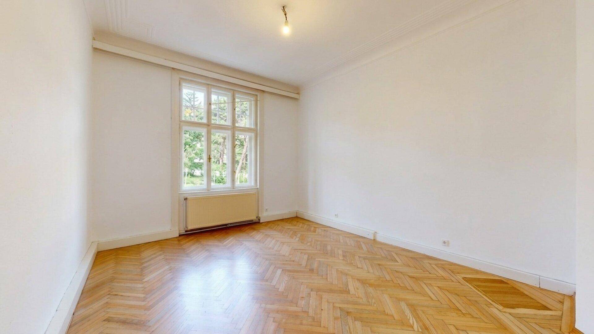 Helles, leeres Zimmer mit Fischgrätparkett und einem großen Fenster, das viel Licht spendet.