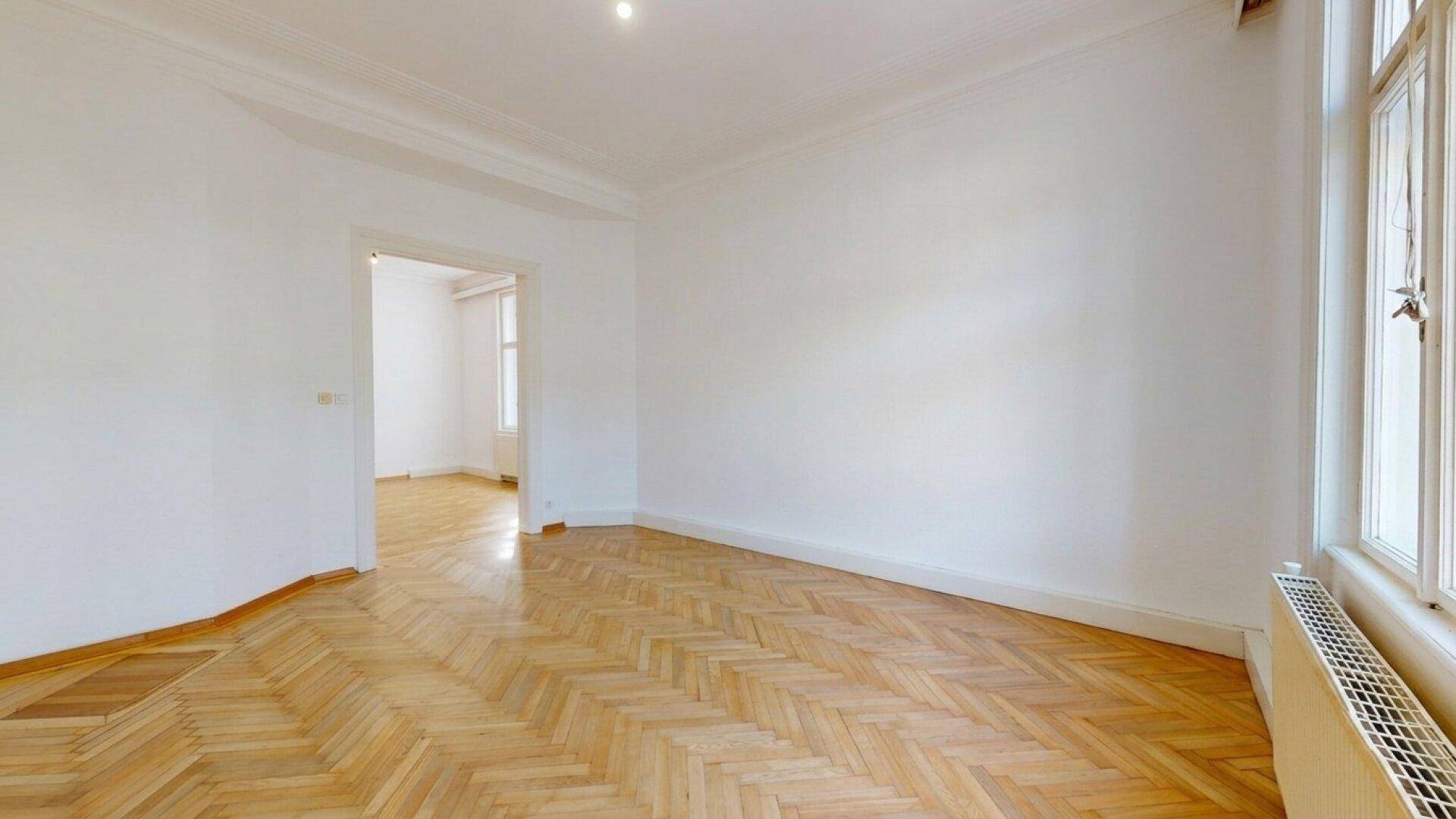 Leeres Zimmer mit Fischgrätparkett und hohen Decken, das durch eine Doppeltür mit einem weiteren Raum verbunden ist.