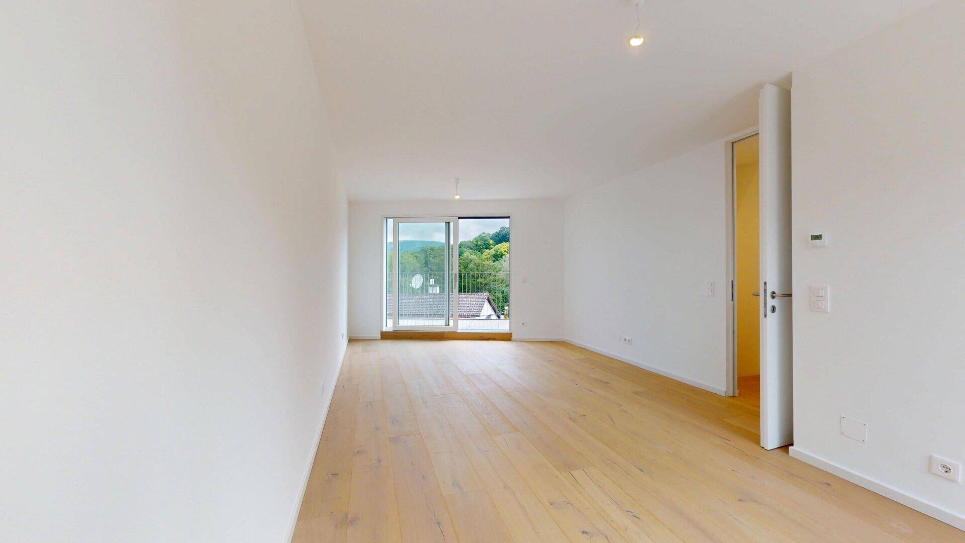 Helles Zimmer mit Holzboden und Zugang zu einem Balkon mit Blick ins Grüne.