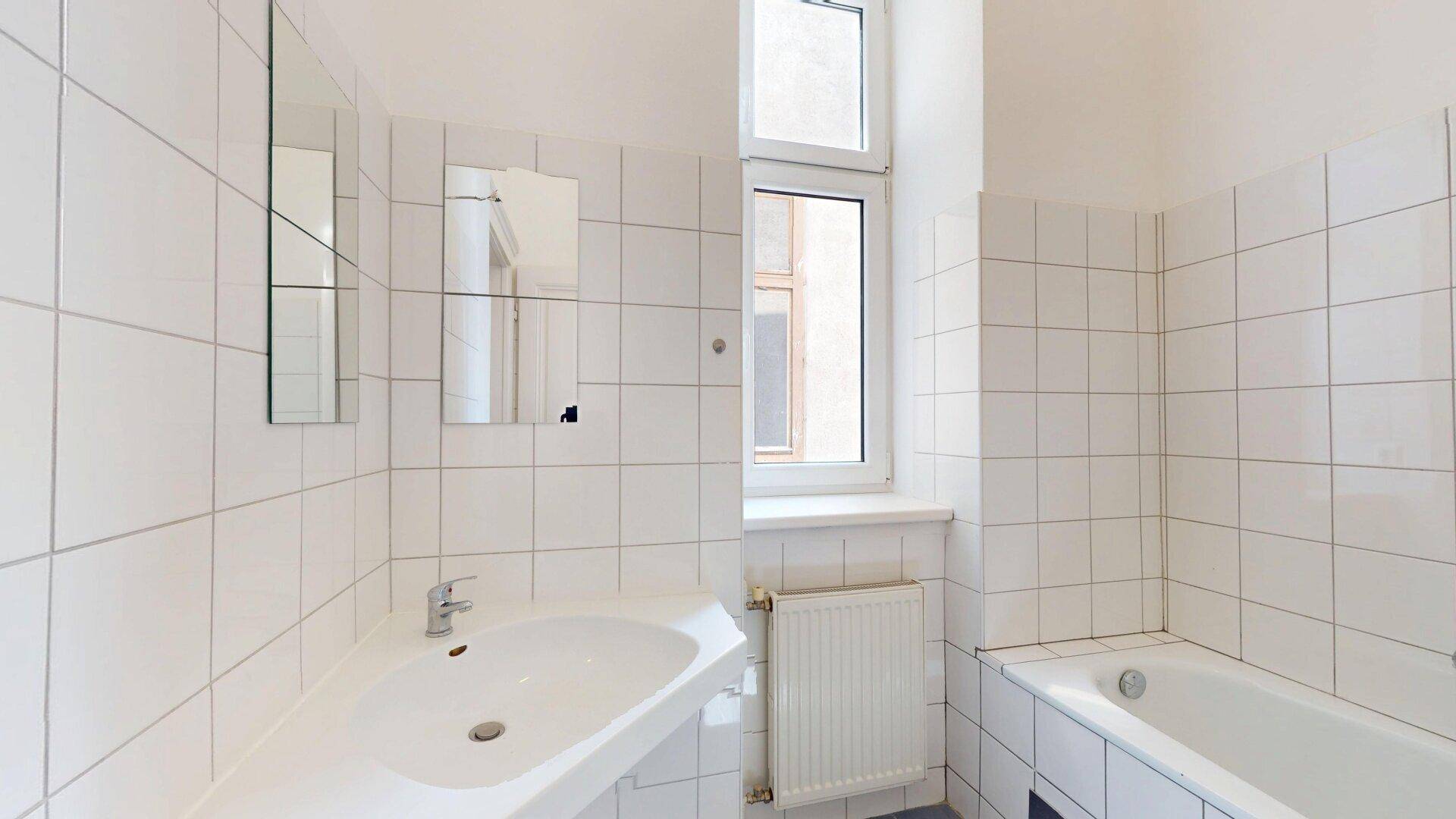 Badezimmer mit weißen Fliesen, Waschbecken, Spiegel, Badewanne und Fenster.