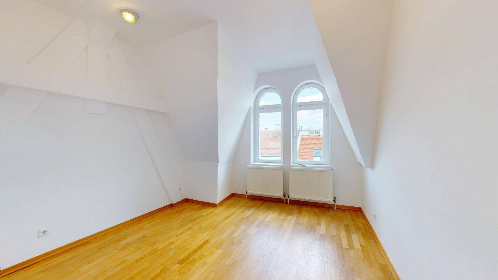 Gemütliches Zimmer im Dachgeschoss mit Holzfußboden, Dachschräge und zwei Rundbogenfenstern.