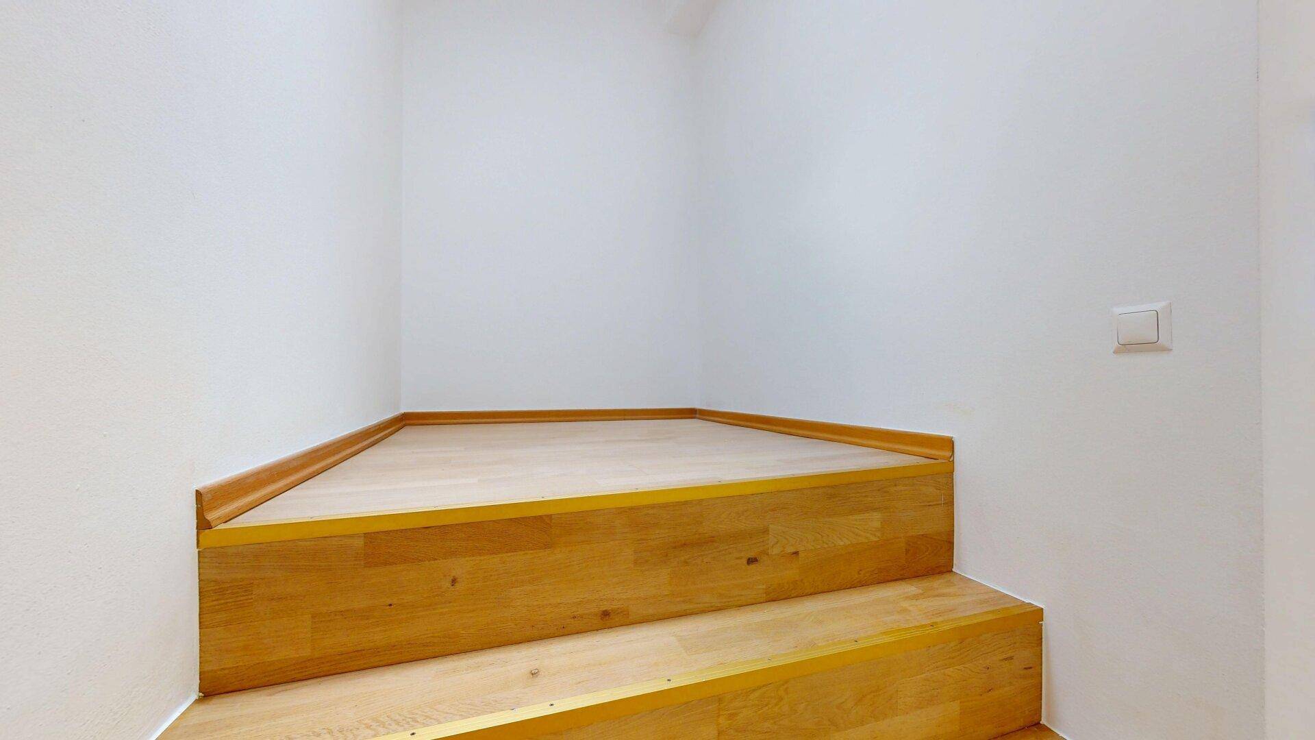 Holztreppe mit hellen Stufen und weiß gestrichenen Wänden, die zum oberen Stockwerk führt.