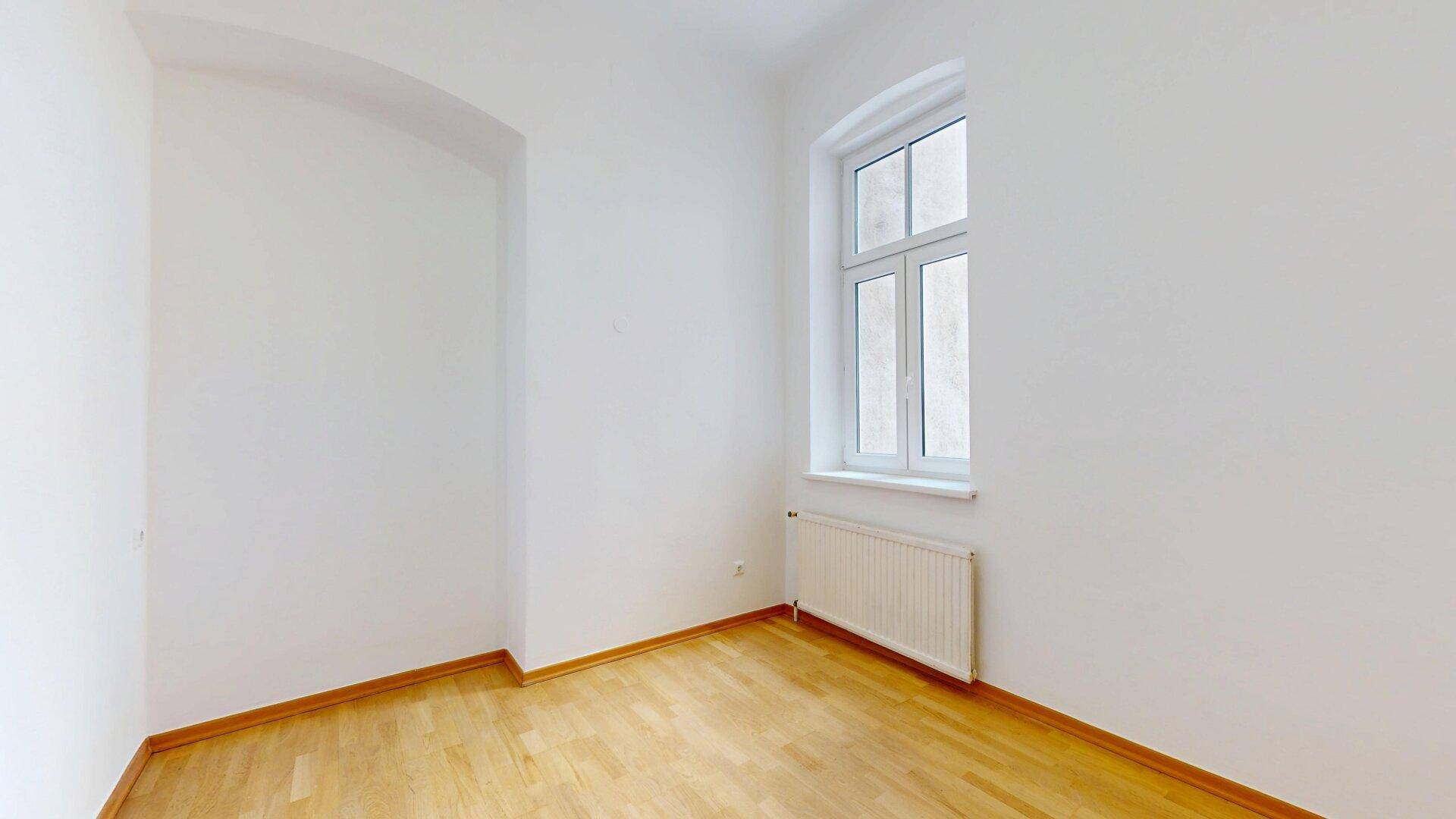 Helles Zimmer mit Holzfußboden, weißen Wänden und einem Fenster mit Blick nach draußen.