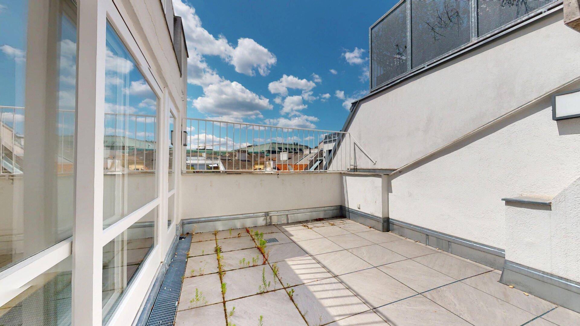 Dachterrasse mit Fliesenboden, Metallgeländer und Blick über die Dächer der Stadt.