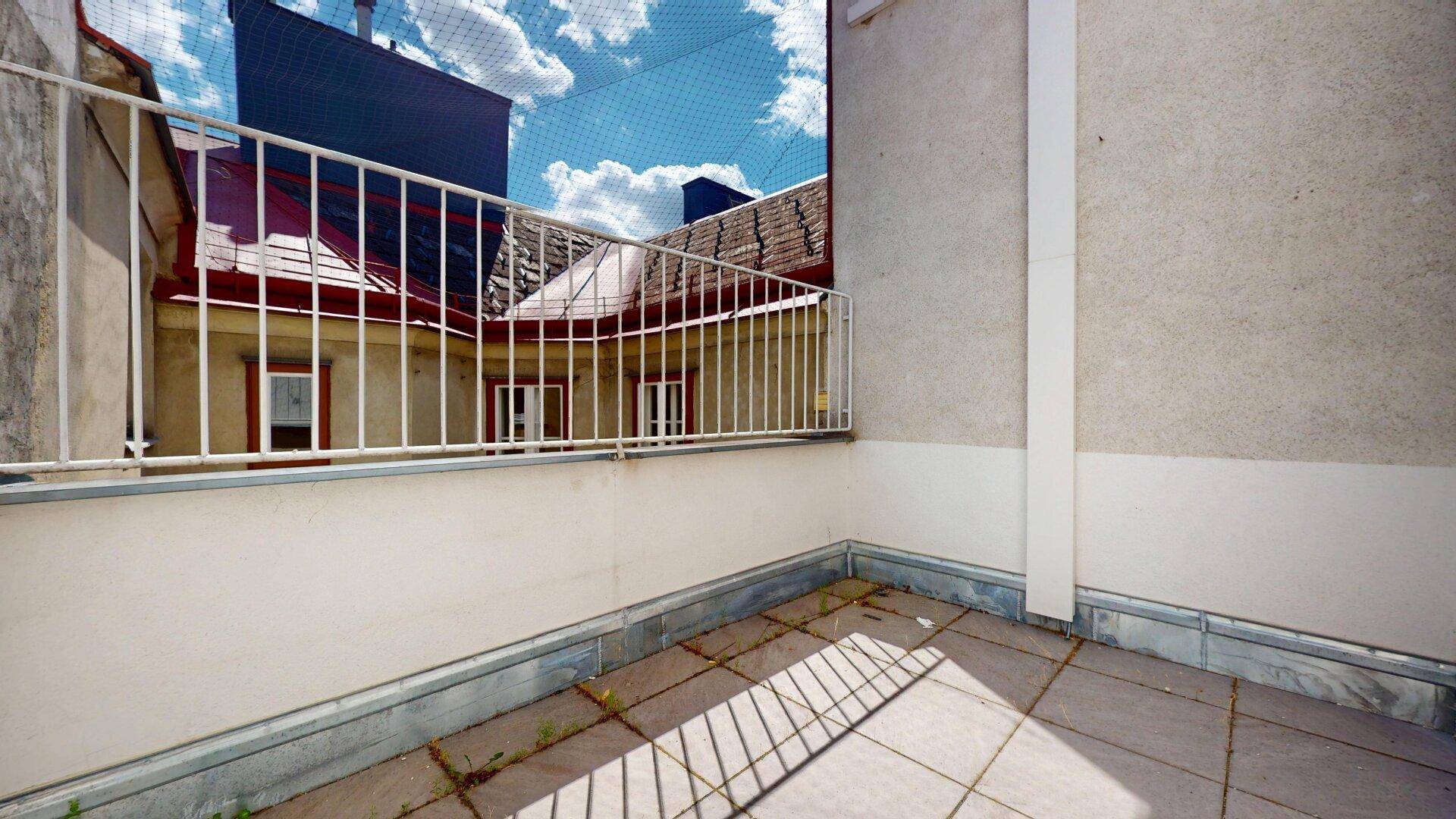 Kleiner Balkon mit Fliesenboden und Blick auf umliegende Gebäude unter einem blauen Himmel.