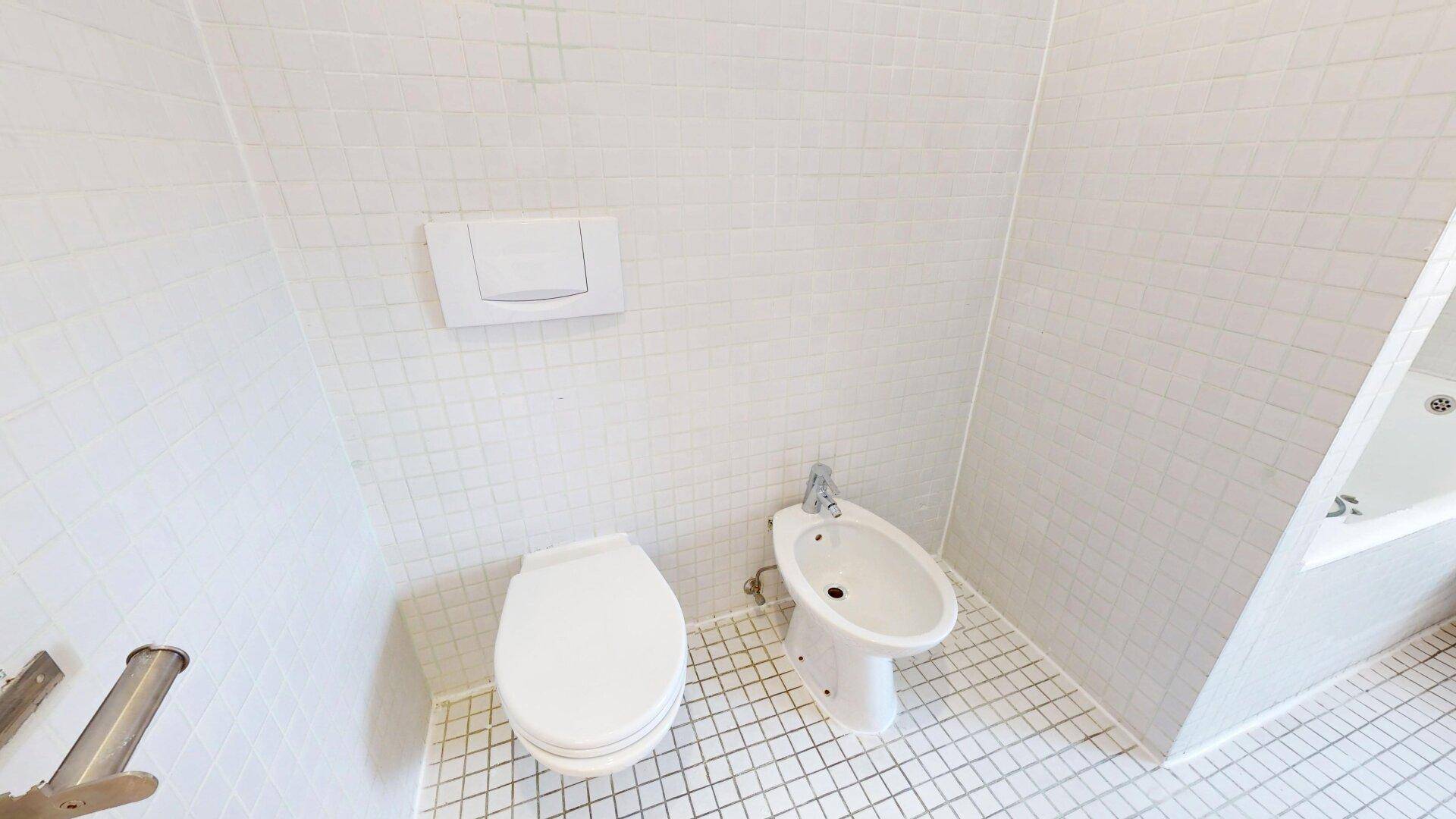 Badezimmer mit Toilette, Bidet und durchgehend weißen Fliesen an Wänden und Boden.