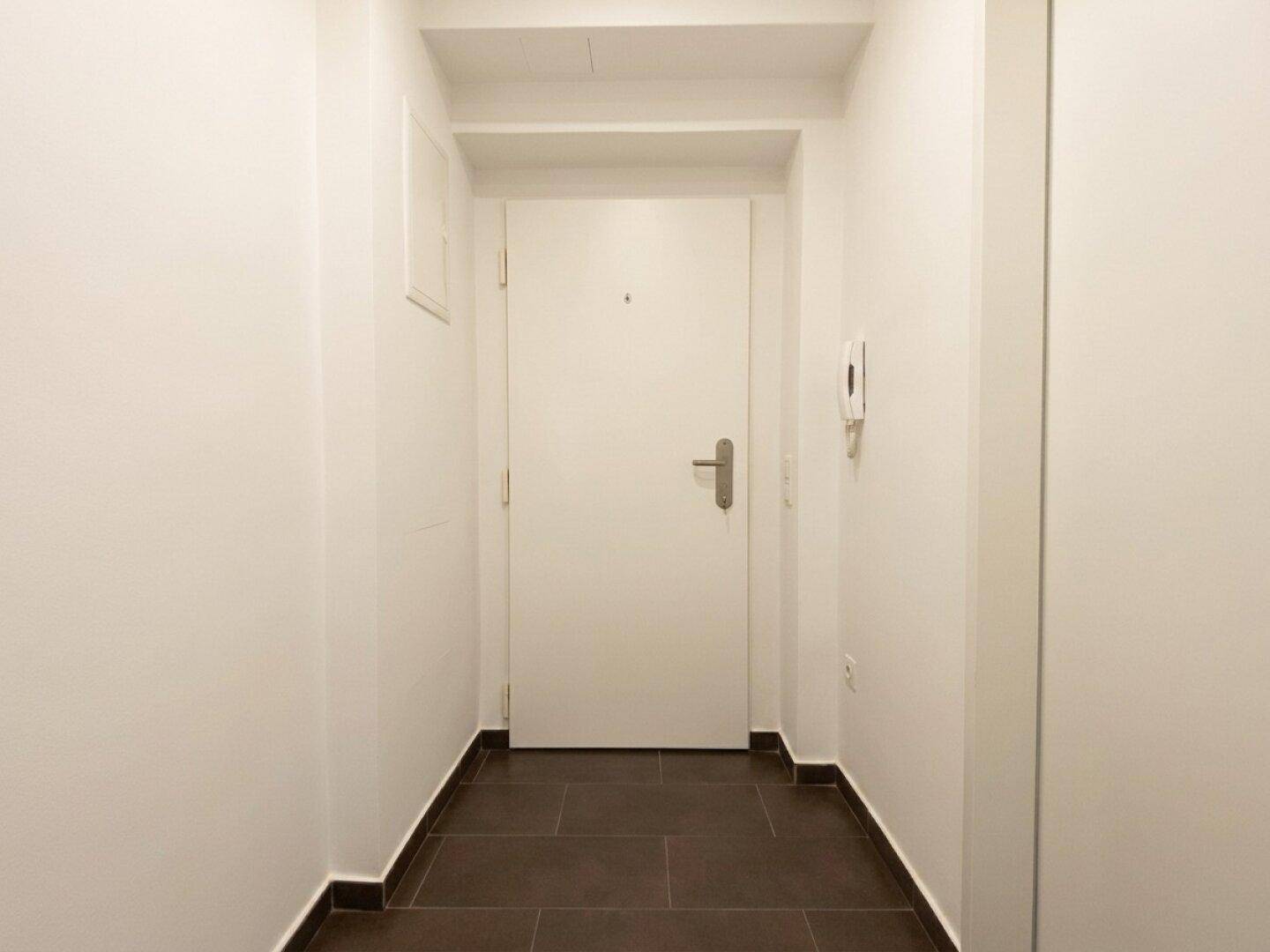 Heller Flur mit dunklen Fliesen und weißer Eingangstür, die zu den Wohnräumen führt.