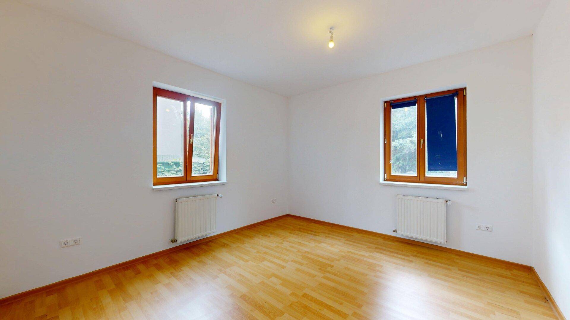 Helles Schlafzimmer mit Laminatboden und zwei Fenstern mit Blick ins Grüne.