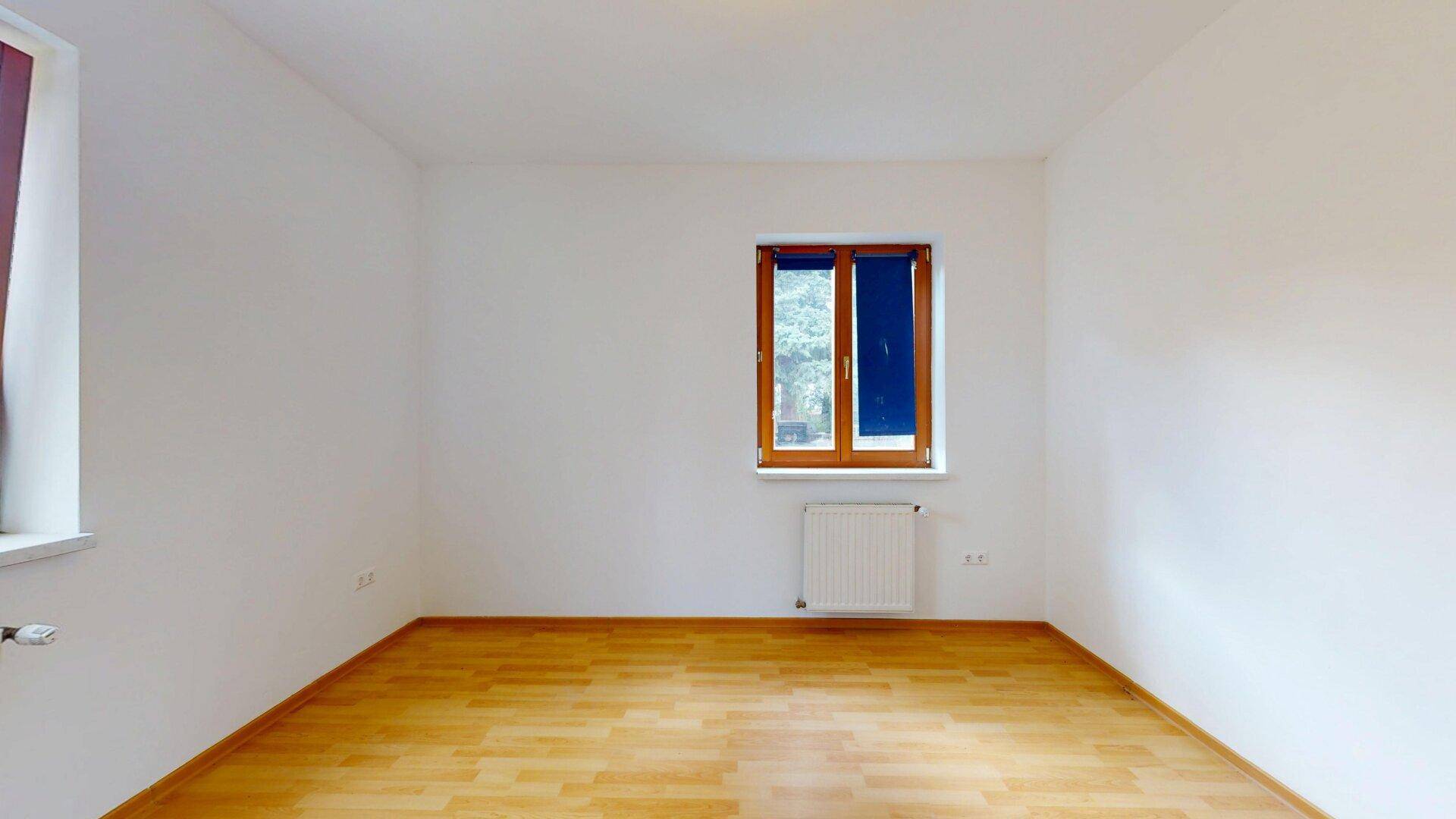 Helles, leeres Zimmer mit Parkettboden und einem Fenster mit blauen Rollos.