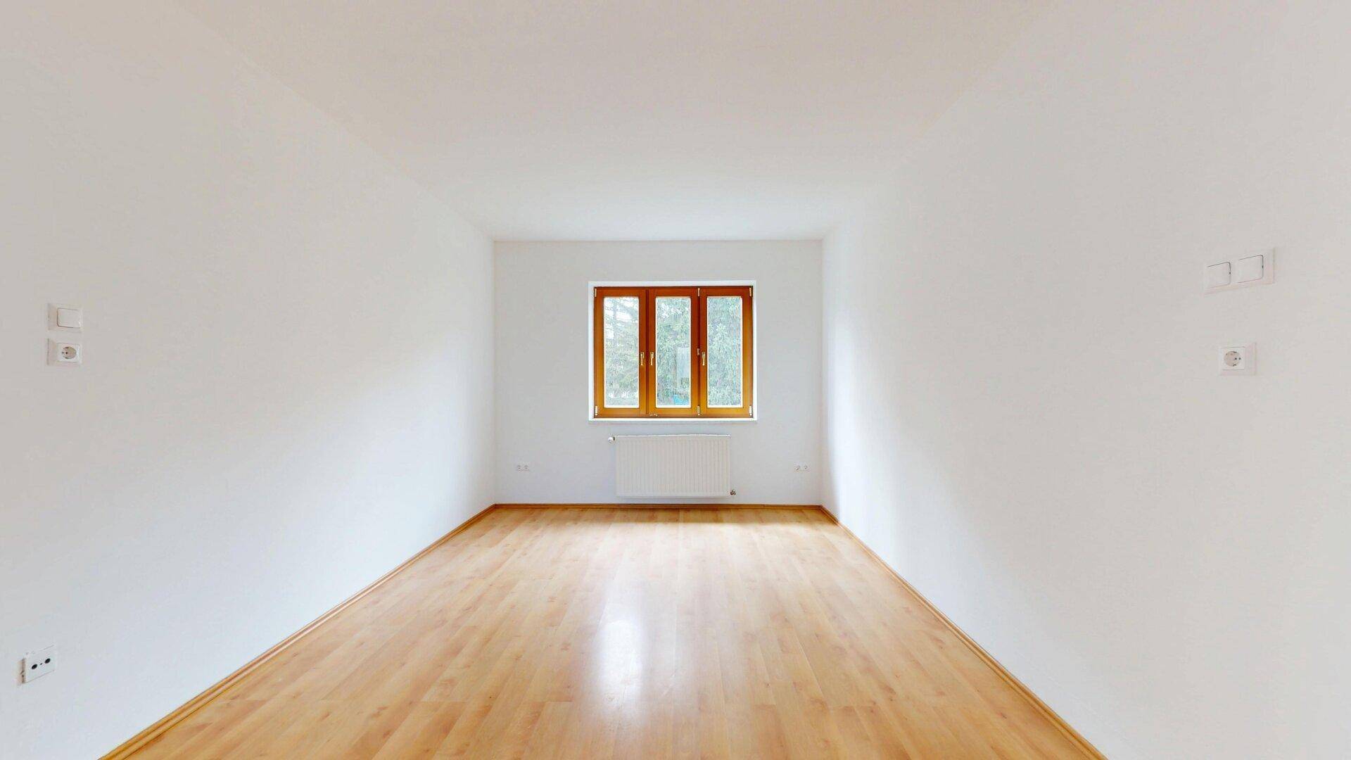 Helles, leeres Zimmer mit Parkettboden und einem breiten Fenster mit Holzrahmen.