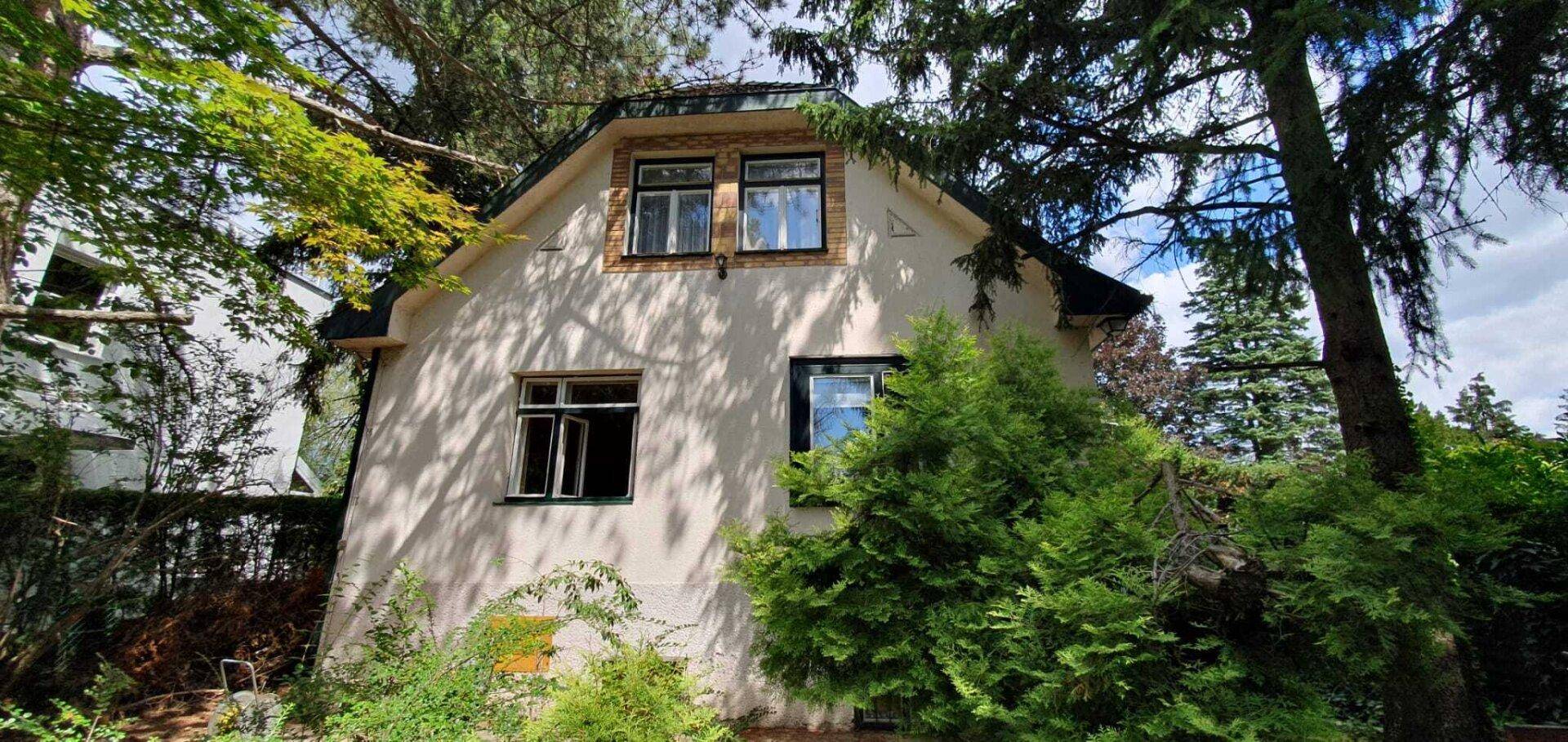 Die Fassade des Hauses ist von Bäumen und Sträuchern umgeben, mit sichtbaren Fenstern und Dach.