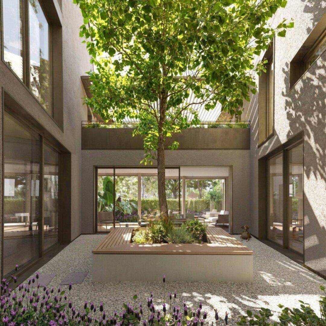 Grüner Innenhof mit zentralem Baum und Sitzgelegenheiten, umgeben von modernen Gebäudefassaden.