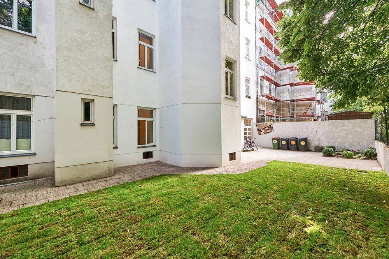 Kleiner Gemeinschaftsgarten mit Rasenfläche und gepflastertem Weg hinter dem Gebäude.