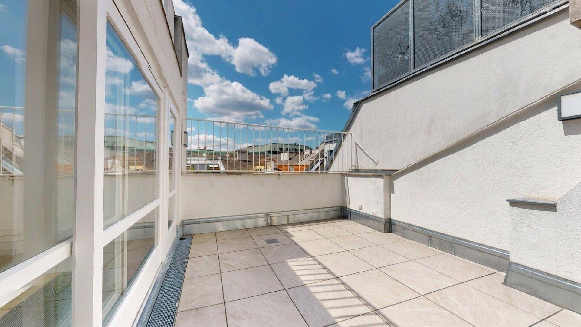 Geräumige Dachterrasse mit hellen Fliesen, weißem Geländer und weitem Blick über die Dächer der Stadt.