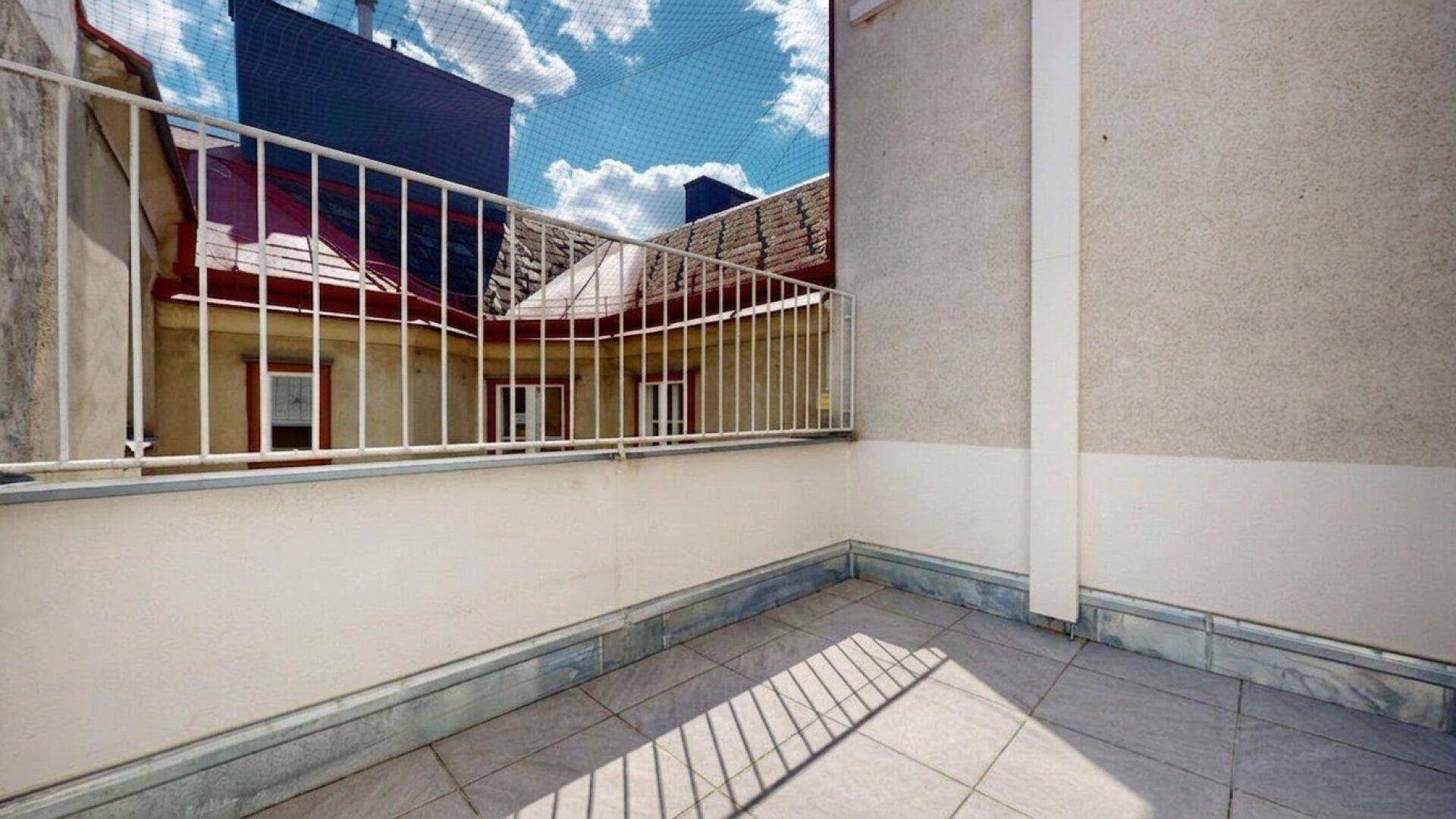 Kleiner Balkon mit grauen Fliesen und weißem Geländer, Blick auf den Innenhof und angrenzende Gebäude.
