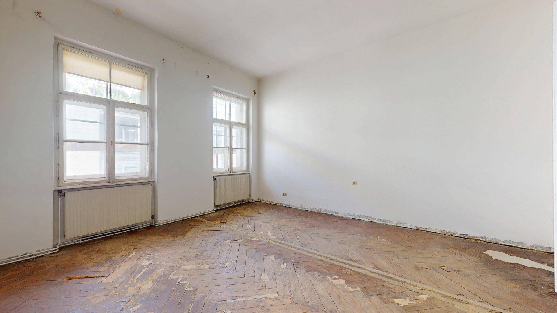 Leeres Zimmer mit abgenutztem Fischgrätparkett und zwei Fenstern, wartet auf Modernisierung.