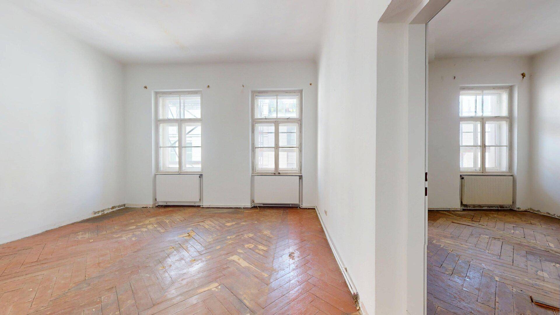 Helles Zimmer mit renovierungsbedürftigem Fischgrätparkett und zwei großen Fenstern.