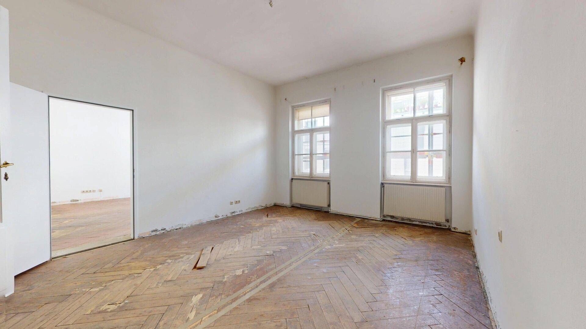 Geräumiges Zimmer mit altem Fischgrätparkett und hellen Fenstern, bereit zur Renovierung.