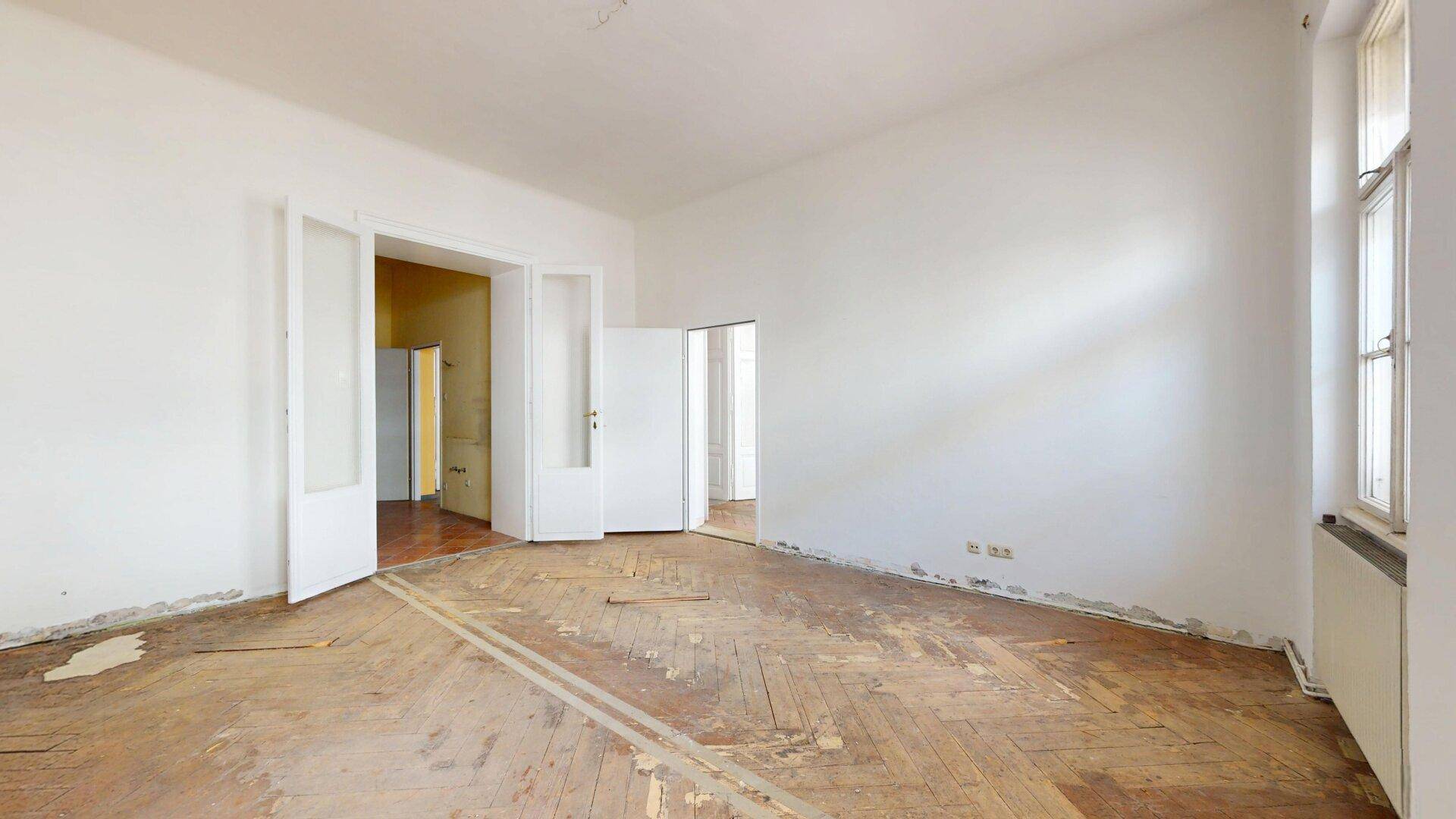 Großes Zimmer mit abgenutztem Fischgrätparkett und doppelten Flügeltüren, ideal für eine Renovierung.