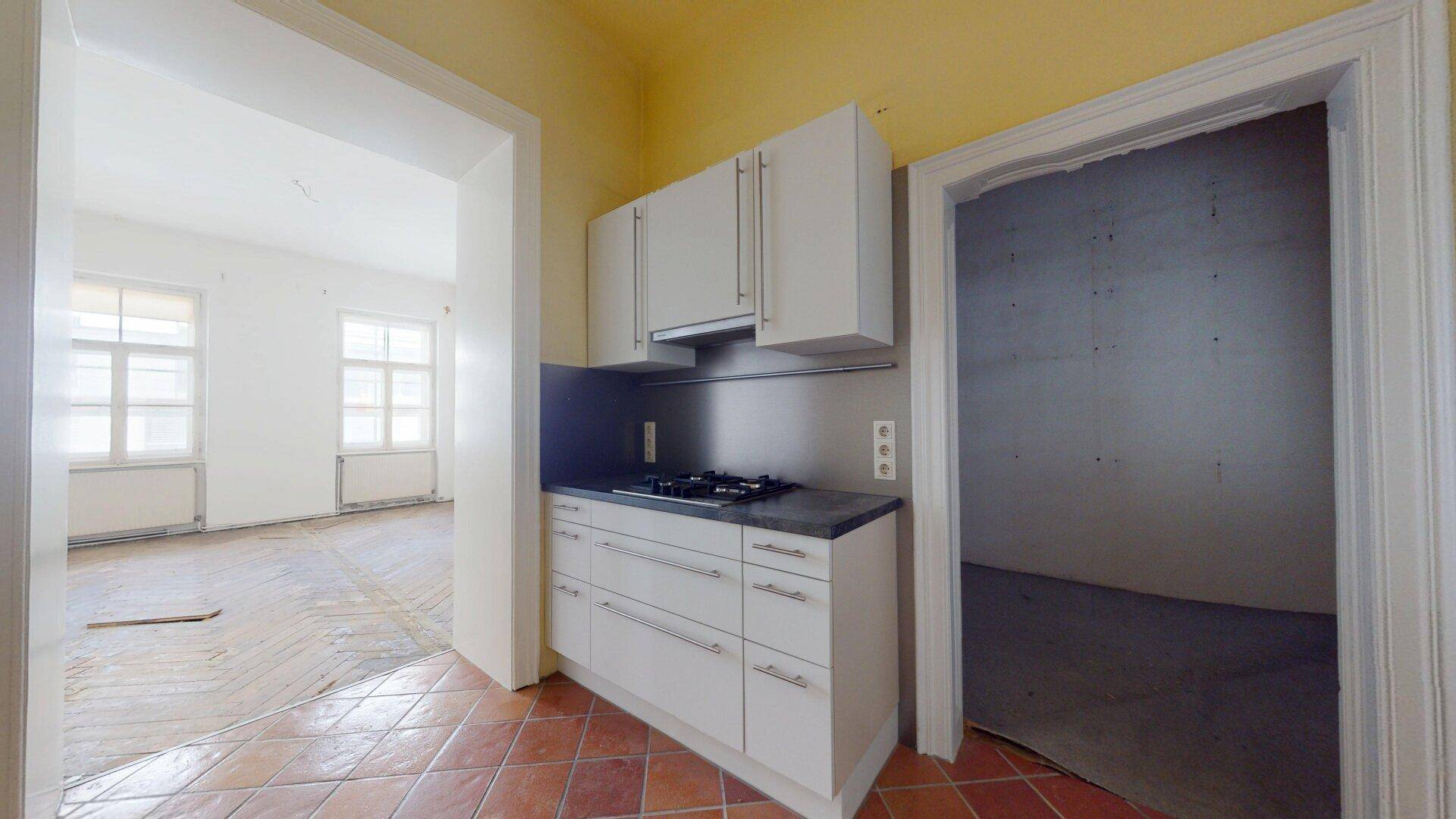 Kleine Küche mit weißen Unterschränken und Gasherd, angrenzend an ein renovierungsbedürftiges Zimmer.