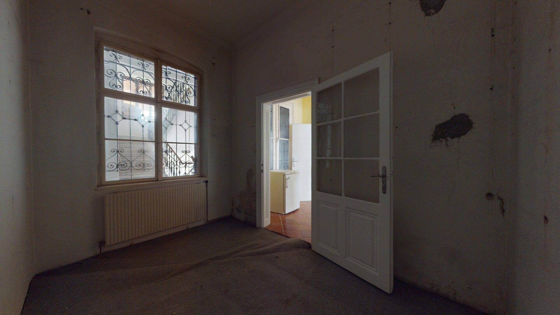 Dunkles Zimmer mit Gitterfenster und renovierungsbedürftigen Wänden, mit Blick auf den Innenhof.