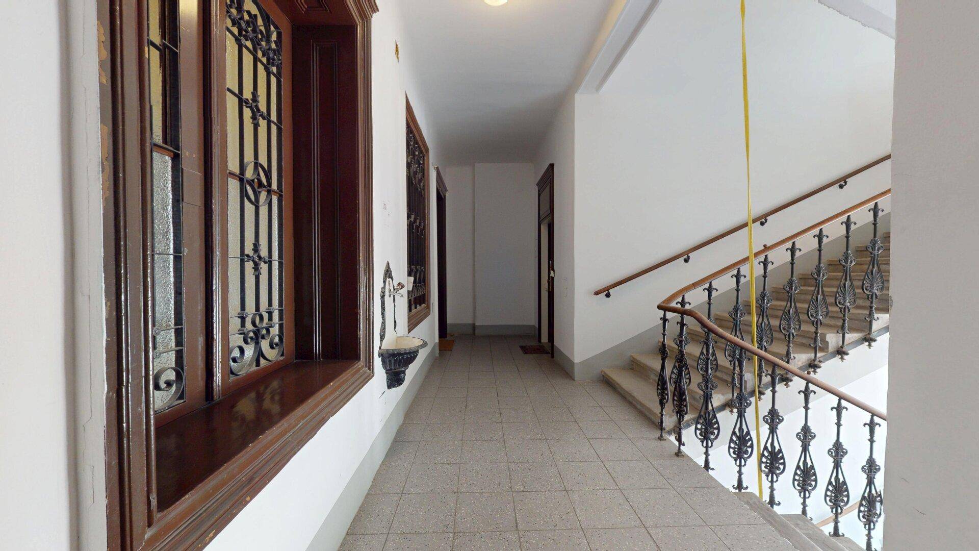 Historisches Treppenhaus mit schmiedeeisernem Geländer und großen Fenstern.