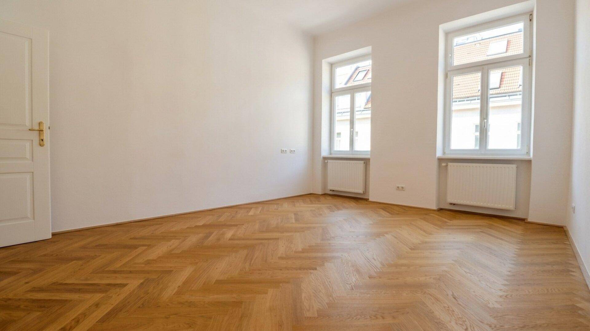 Helles, leeres Zimmer mit Fischgrätparkettboden und zwei großen Fenstern, die viel Tageslicht hereinlassen.