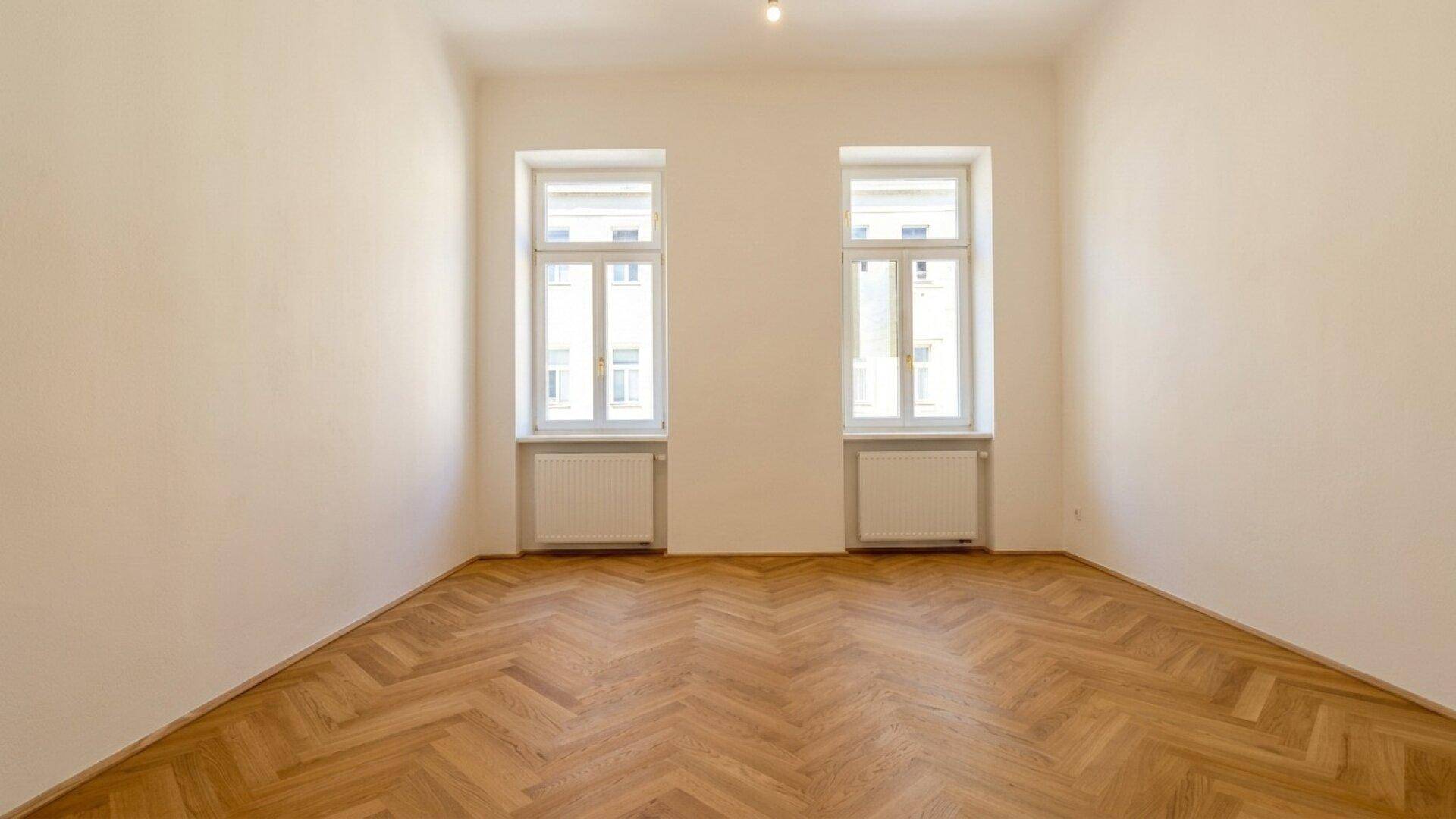 Geräumiges, helles Zimmer mit klassischem Fischgrätparkett und zwei großen Fenstern.